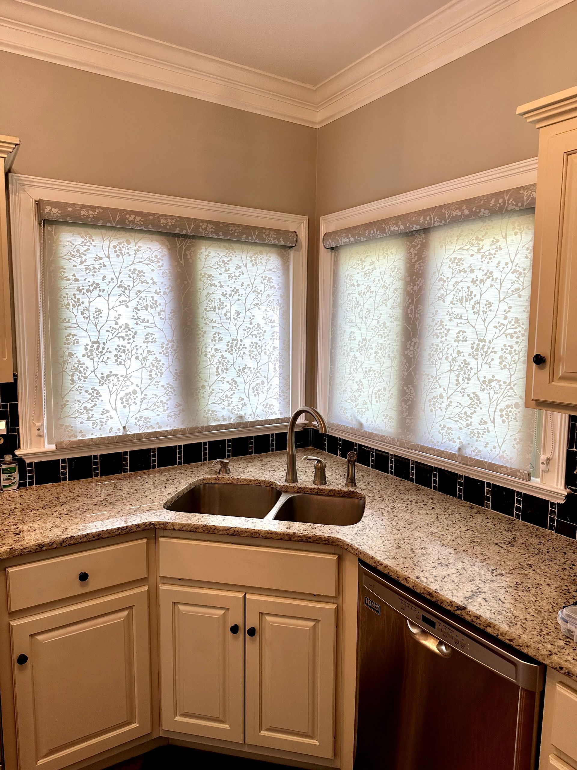 Kitchen corner with a double sink, granite countertop, cream cabinets, and patterned window shades.