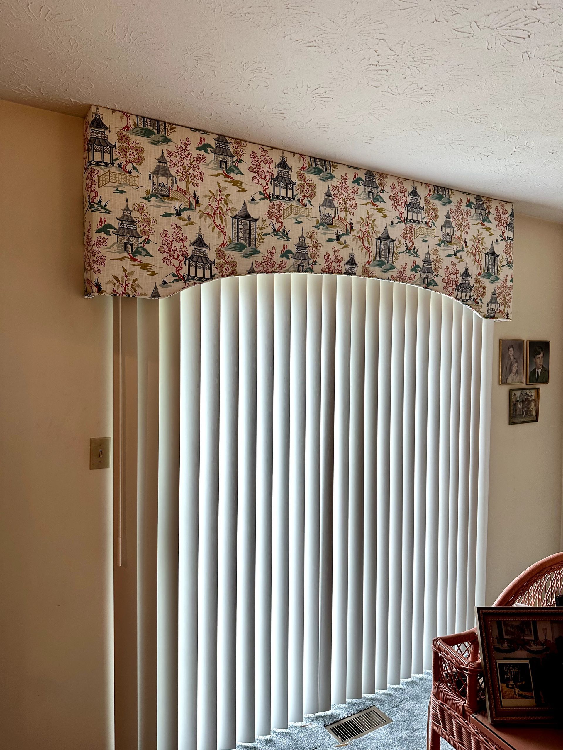 Fabric valance above white vertical blinds, tan walls, and a red wicker chair.