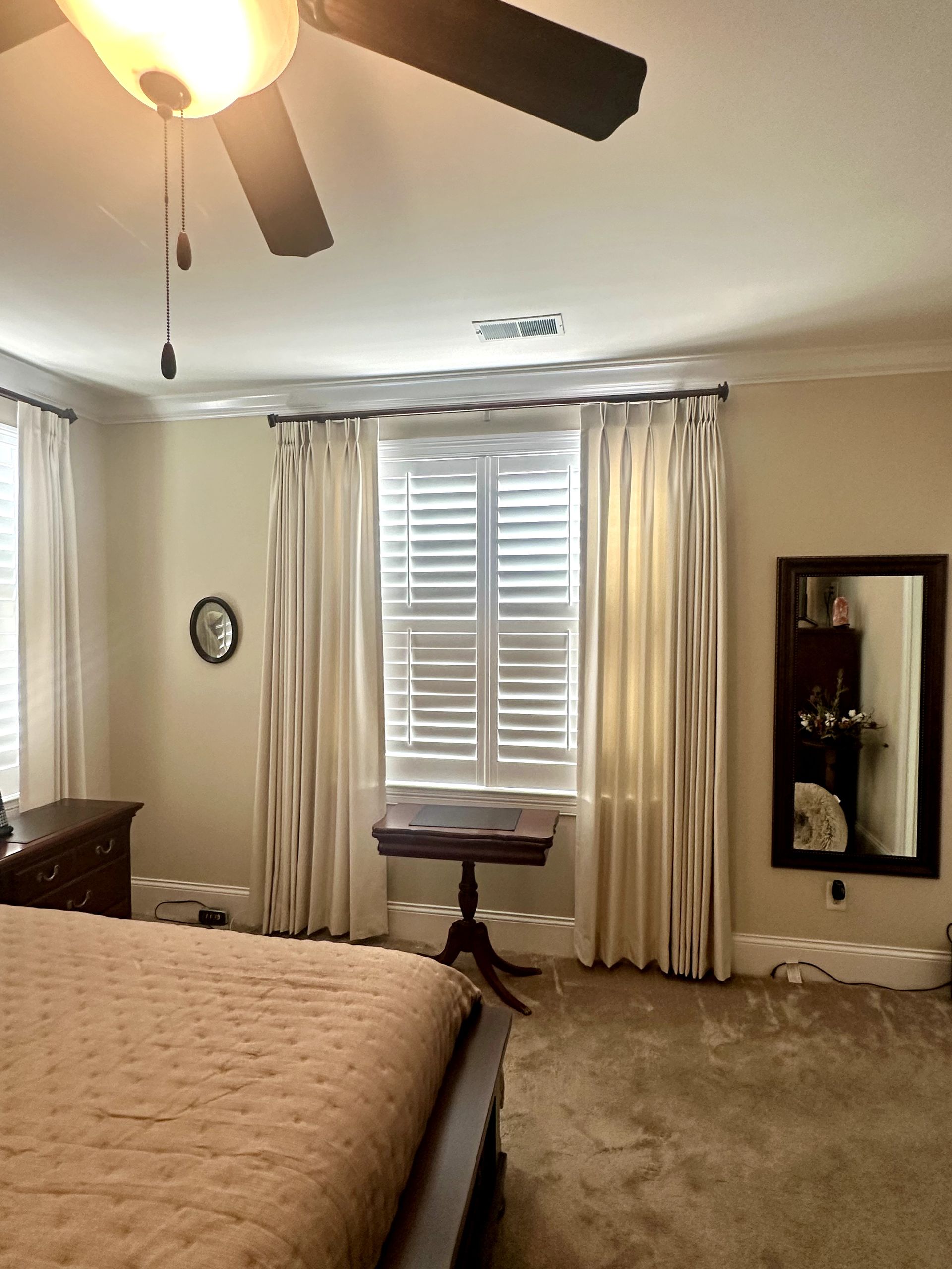 Bedroom with bed, curtains, shutters, table, mirror, and ceiling fan. Beige walls and carpet.