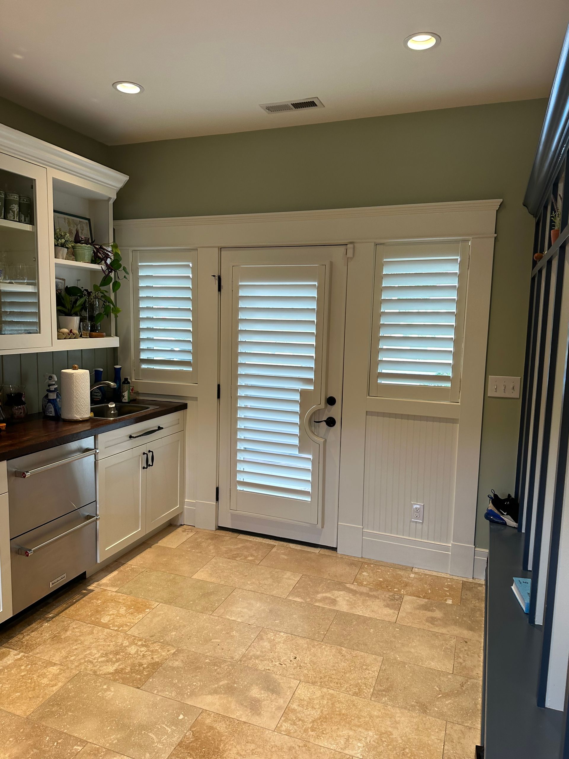 Kitchen with white cabinetry, appliances, and shutters on doors and windows; pale green walls, travertine tile floor.