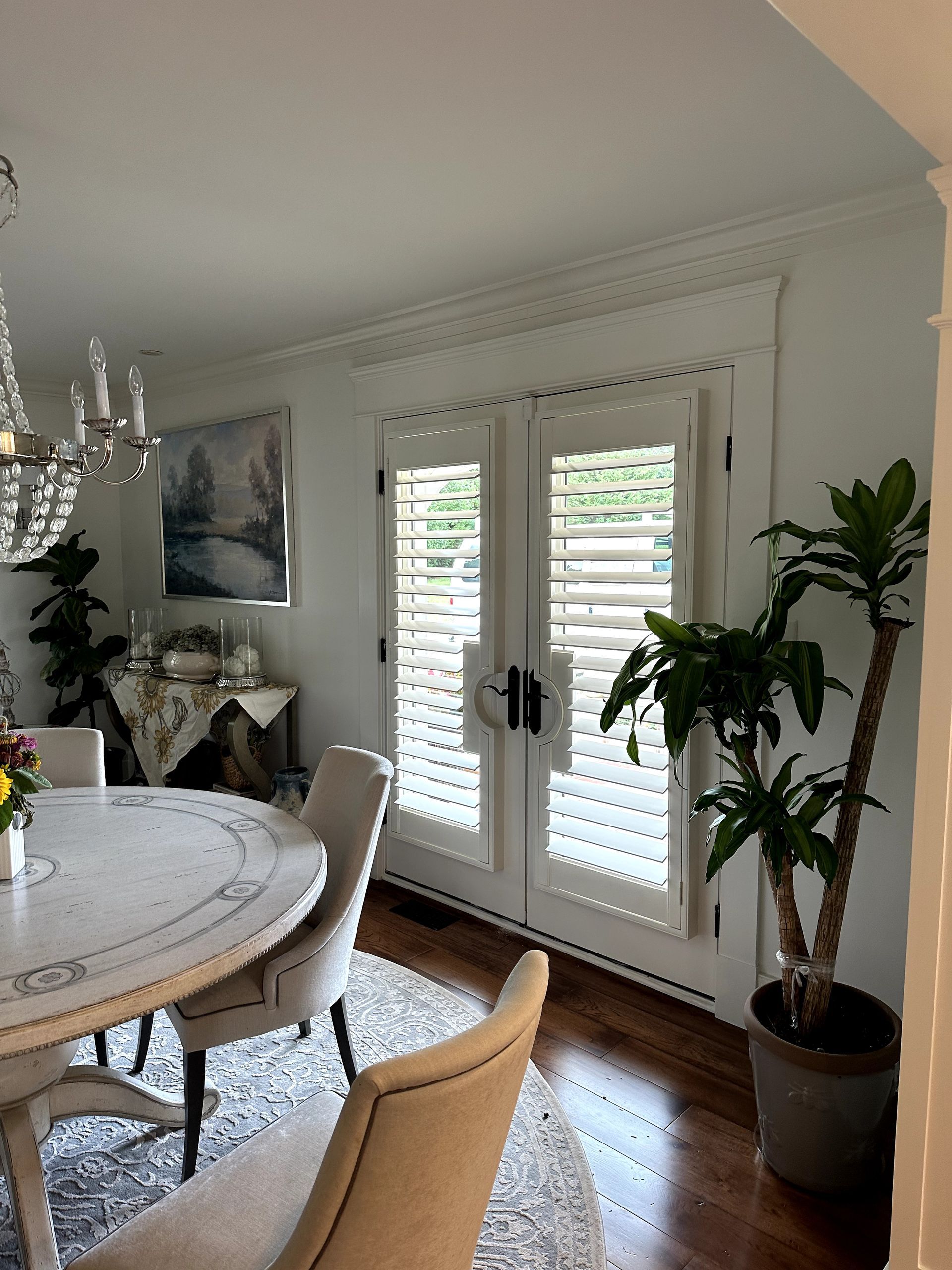 Dining room with round table, chairs, and white shutters on French doors. A chandelier and potted plant are visible.