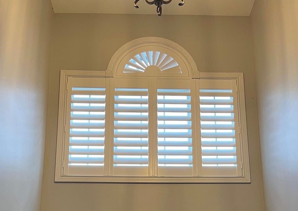 White shutters inside a window frame, with an arched top, set in a light-colored wall.