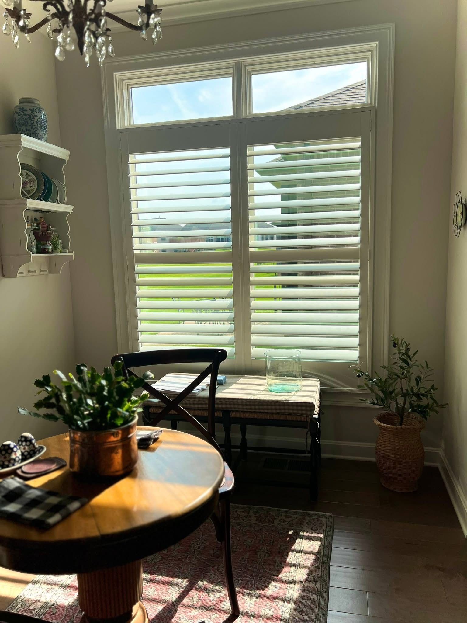 A dining area with a round table, shutters, and plants. Sunlight streams in.