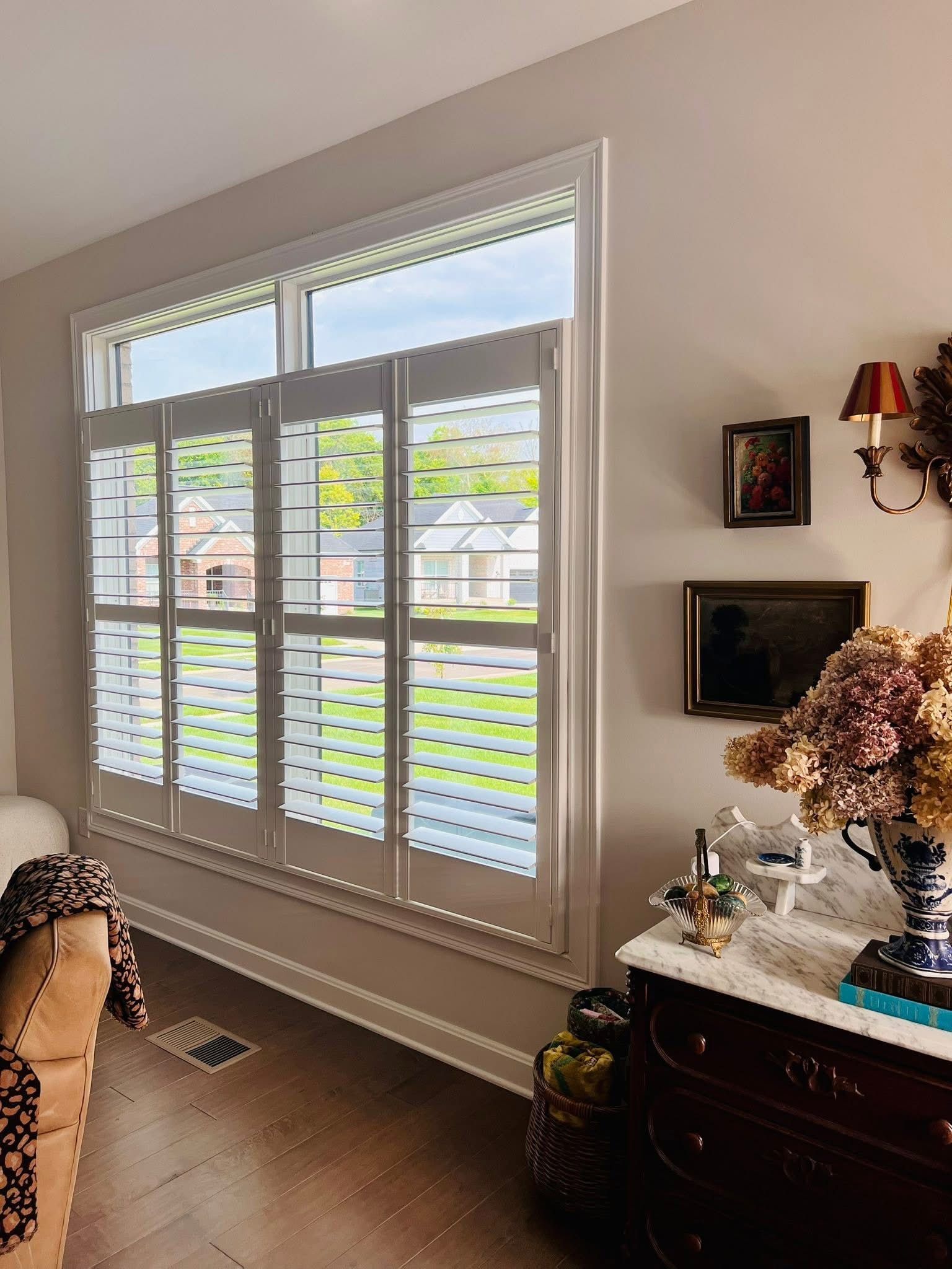 White shutters on a window looking out to a green yard; a brown dresser sits to the right.