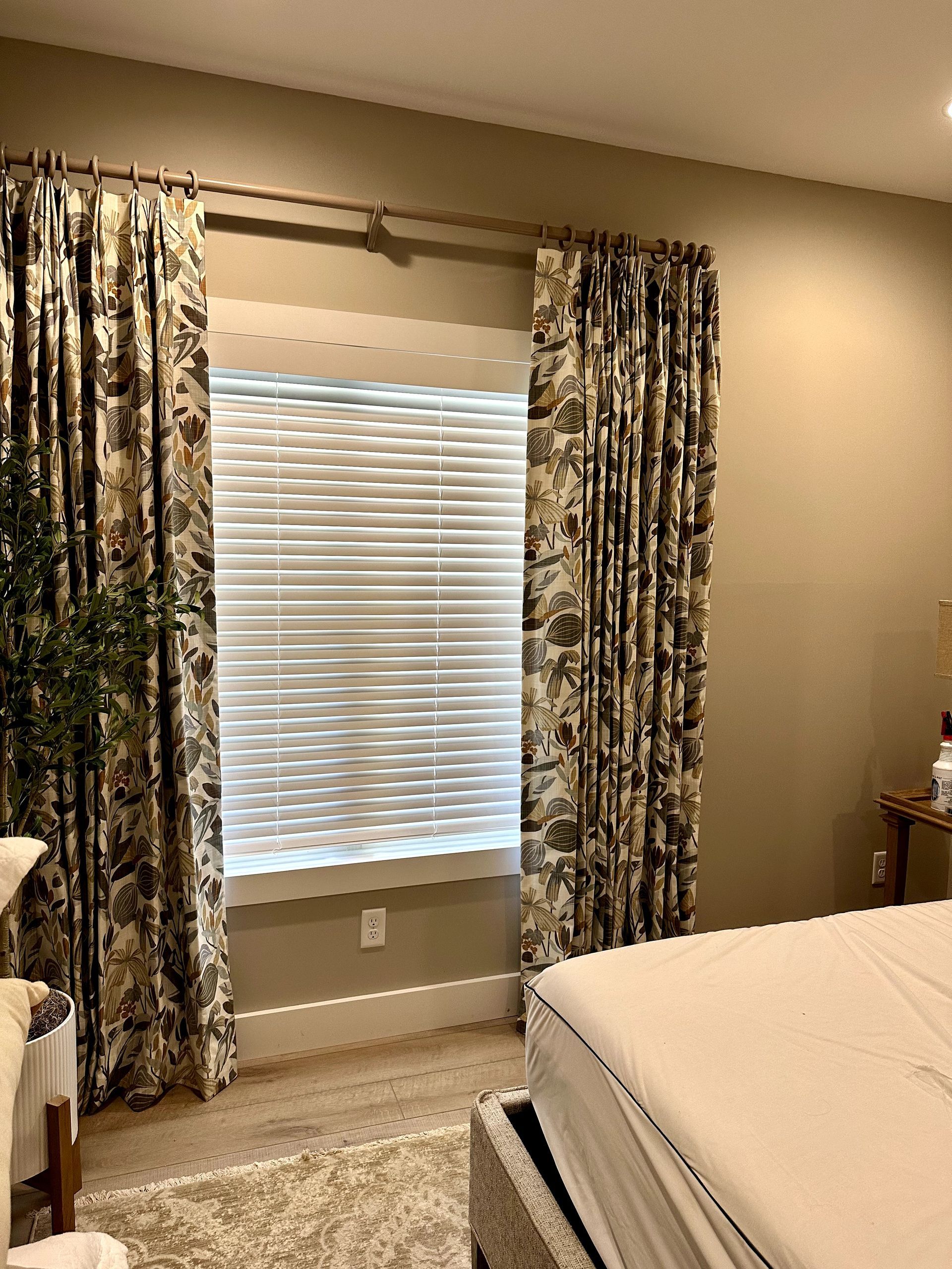 Bedroom with patterned curtains framing a window with white blinds; neutral-toned walls.