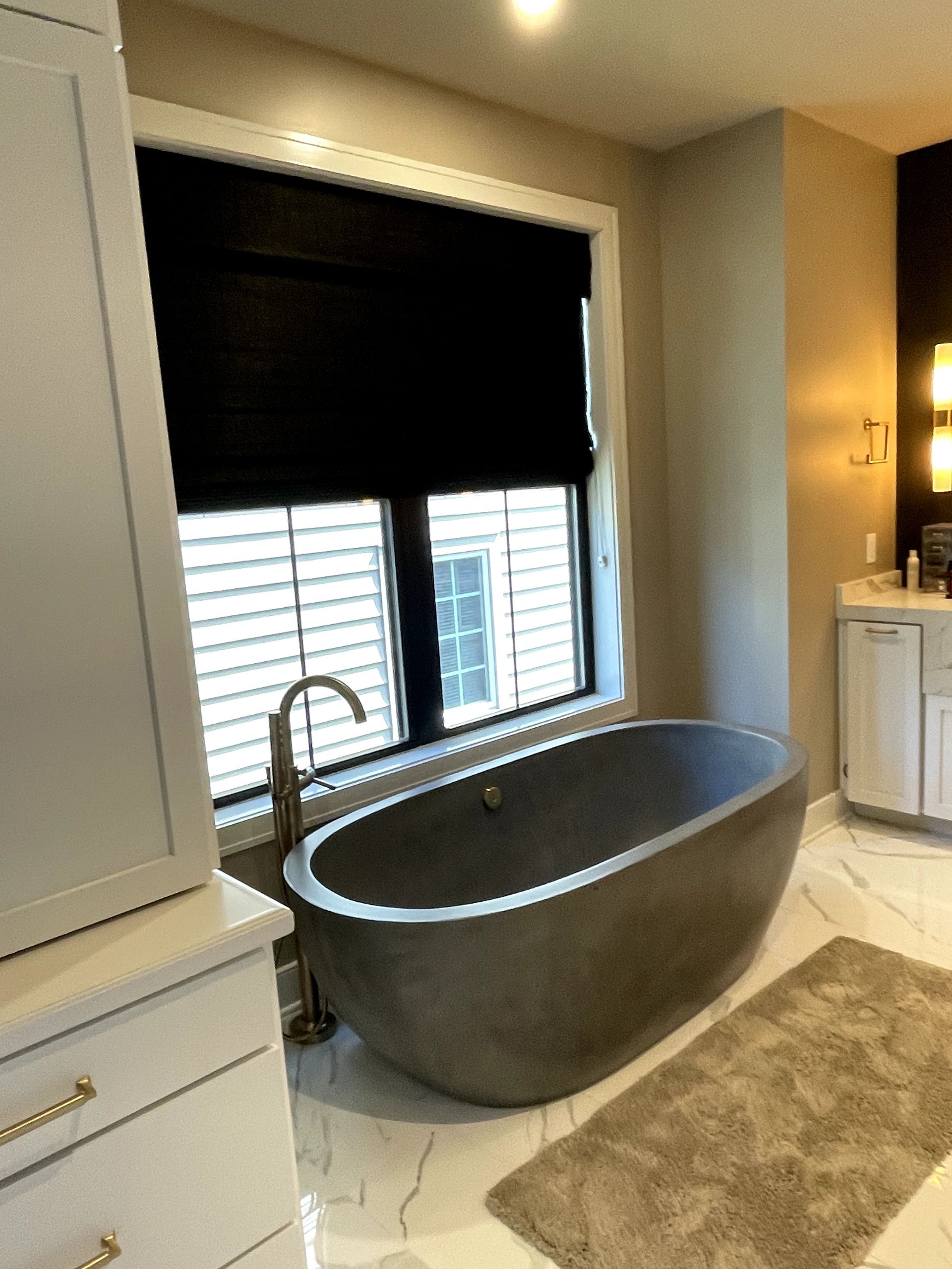 Modern bathroom with gray tub, window with shades, and white cabinets.