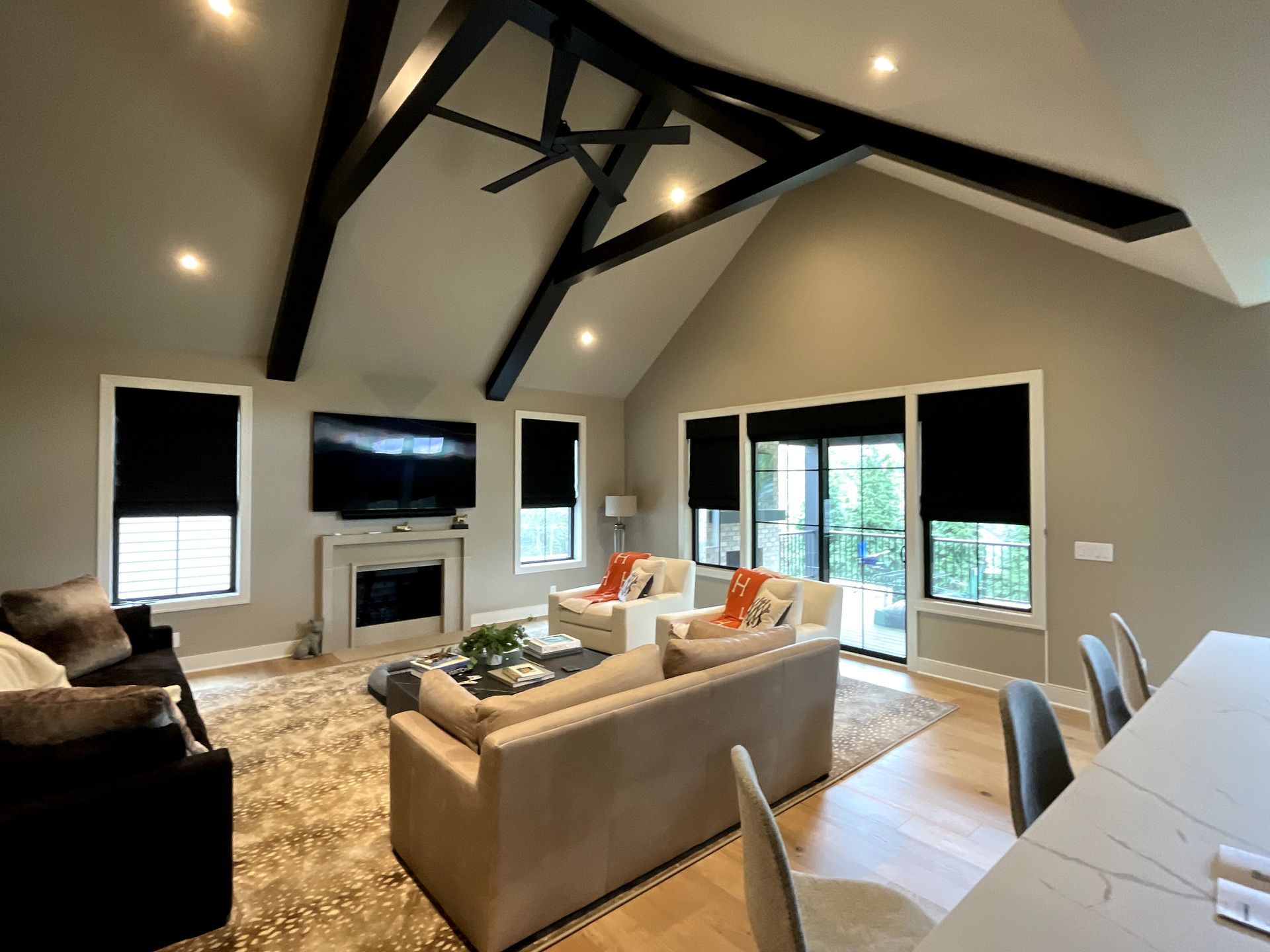 Living room with vaulted ceiling, black beams, fireplace, windows with black shades, and neutral colored furniture.