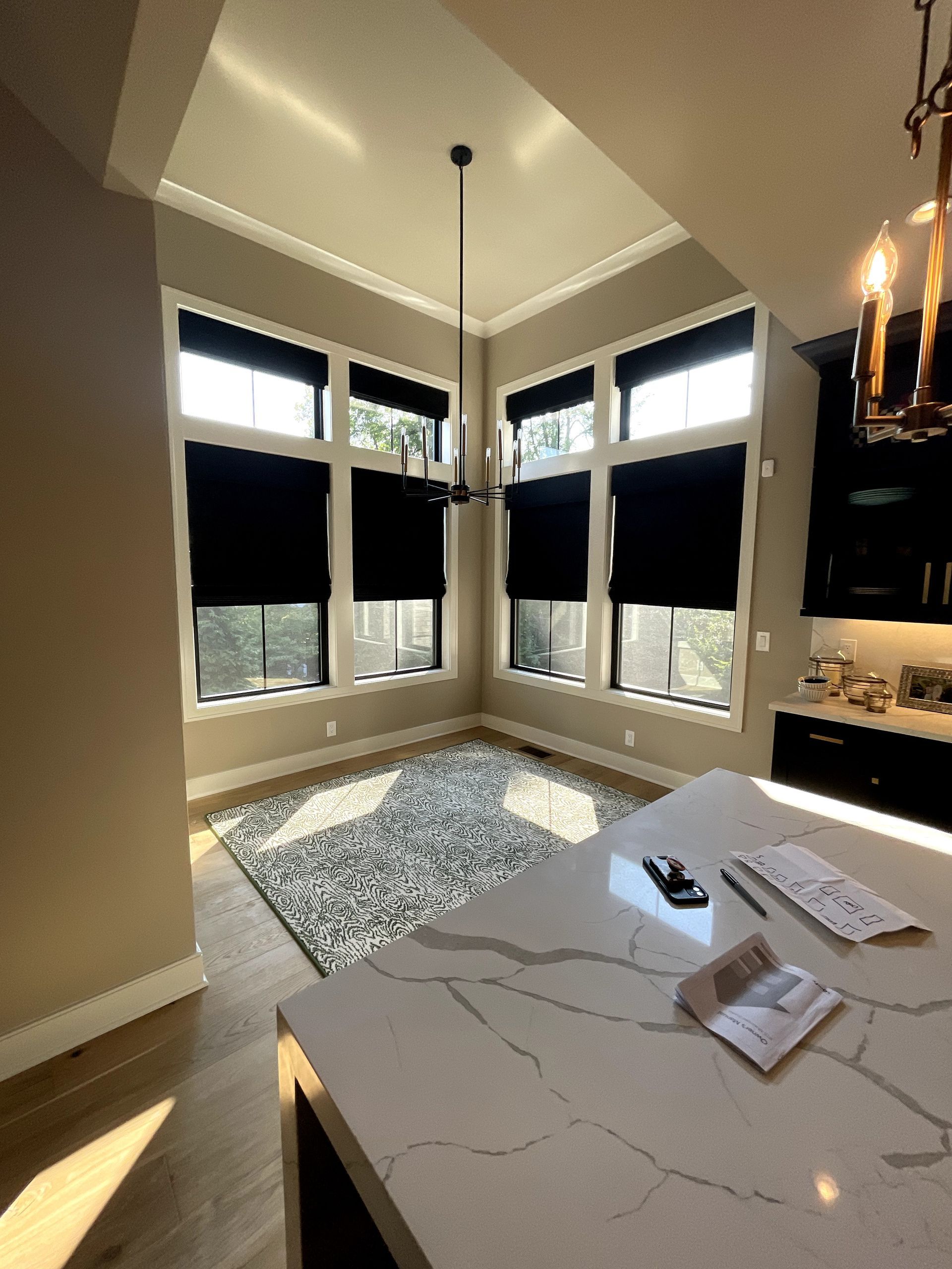 Kitchen with large windows, black shades, chandelier, and white countertop island; natural light streams in.