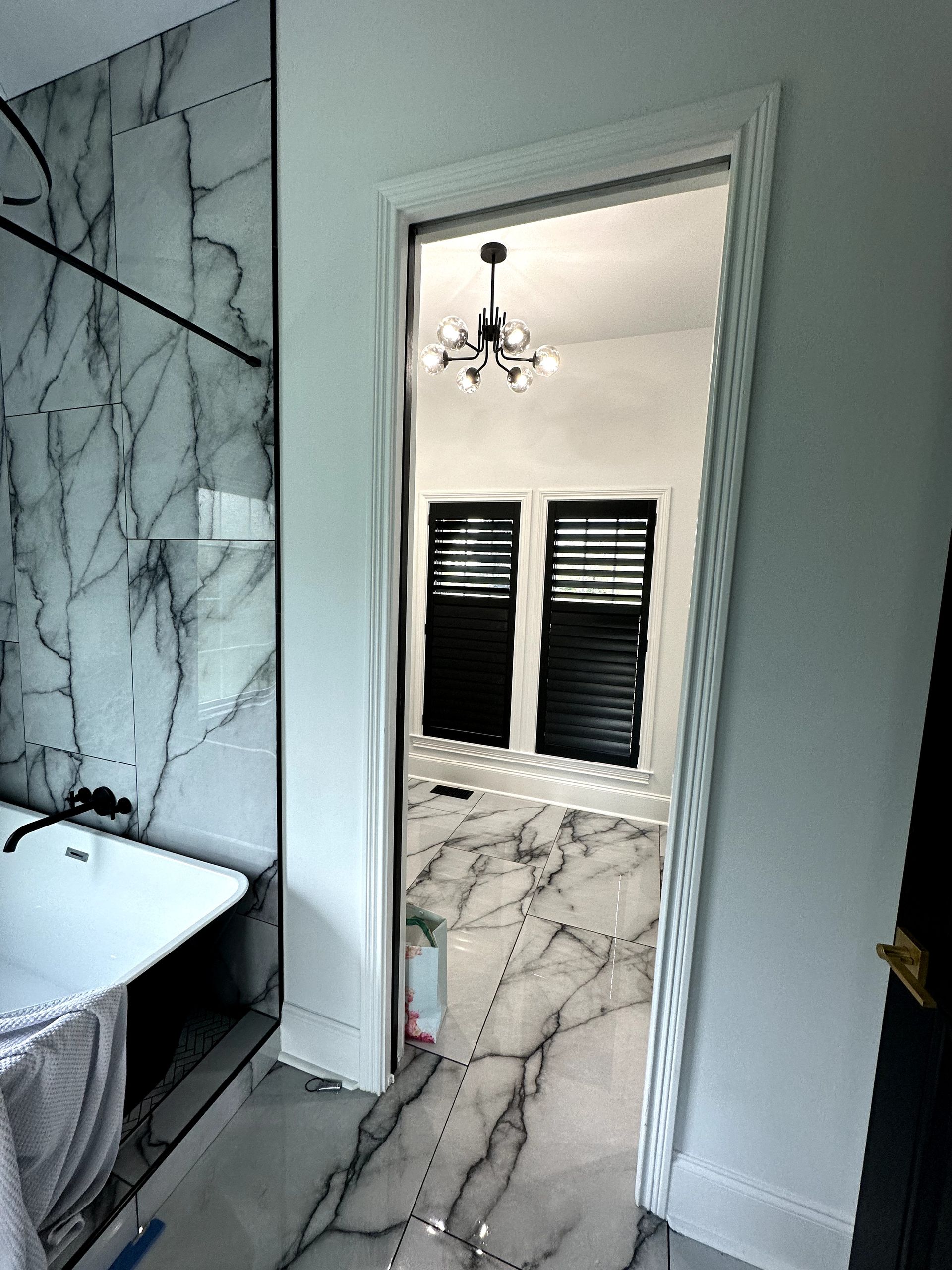 Bathroom with marble tile, open door to a room with black shutters and chandelier.