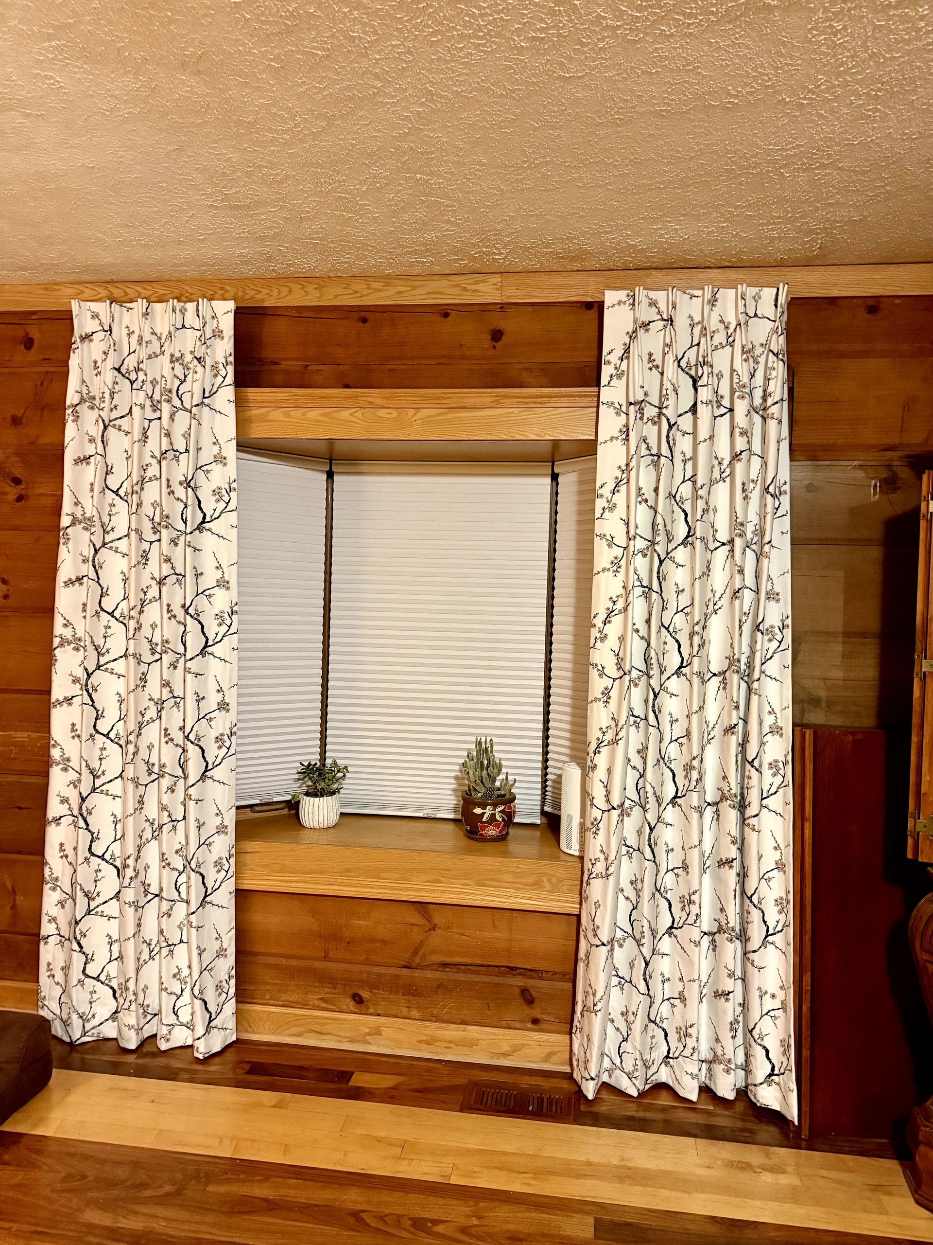 Window with white curtains featuring black floral pattern, set in a wood paneled room.