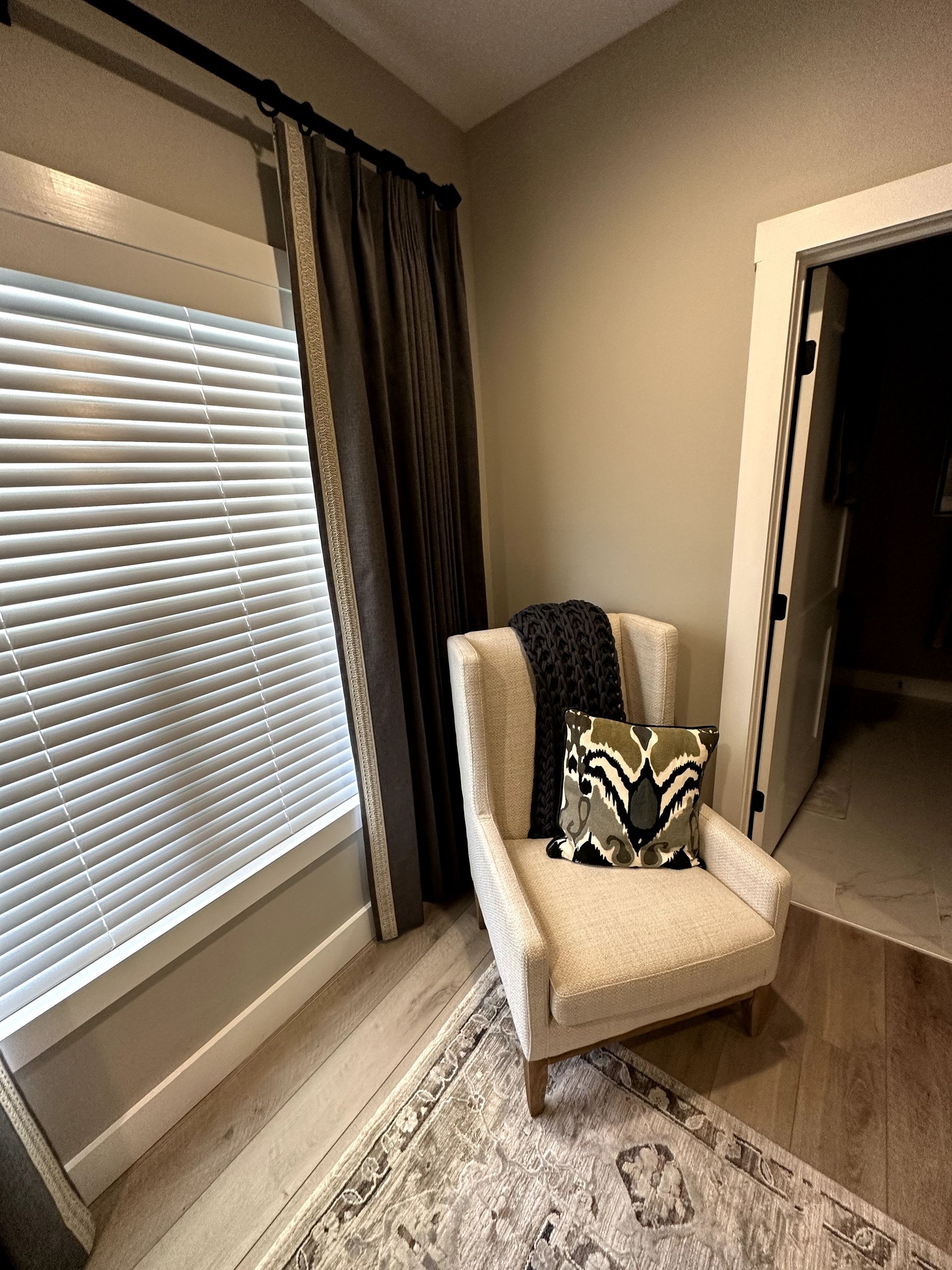 Cozy reading nook: armchair with pillow, dark gray blanket, and patterned rug near a window with blinds.