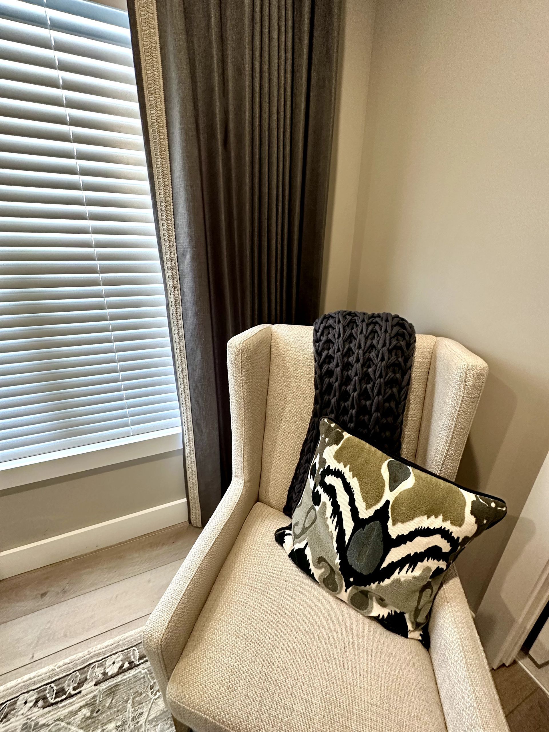 Cozy corner: Cream chair with patterned pillow and blanket, next to a window with blinds and gray curtains.