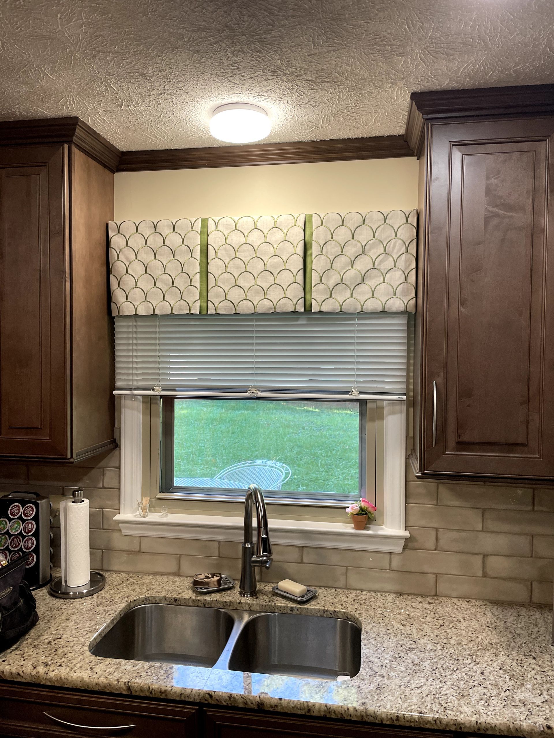 Kitchen with a window, sink, and patterned valance above blinds, and brown cabinets.