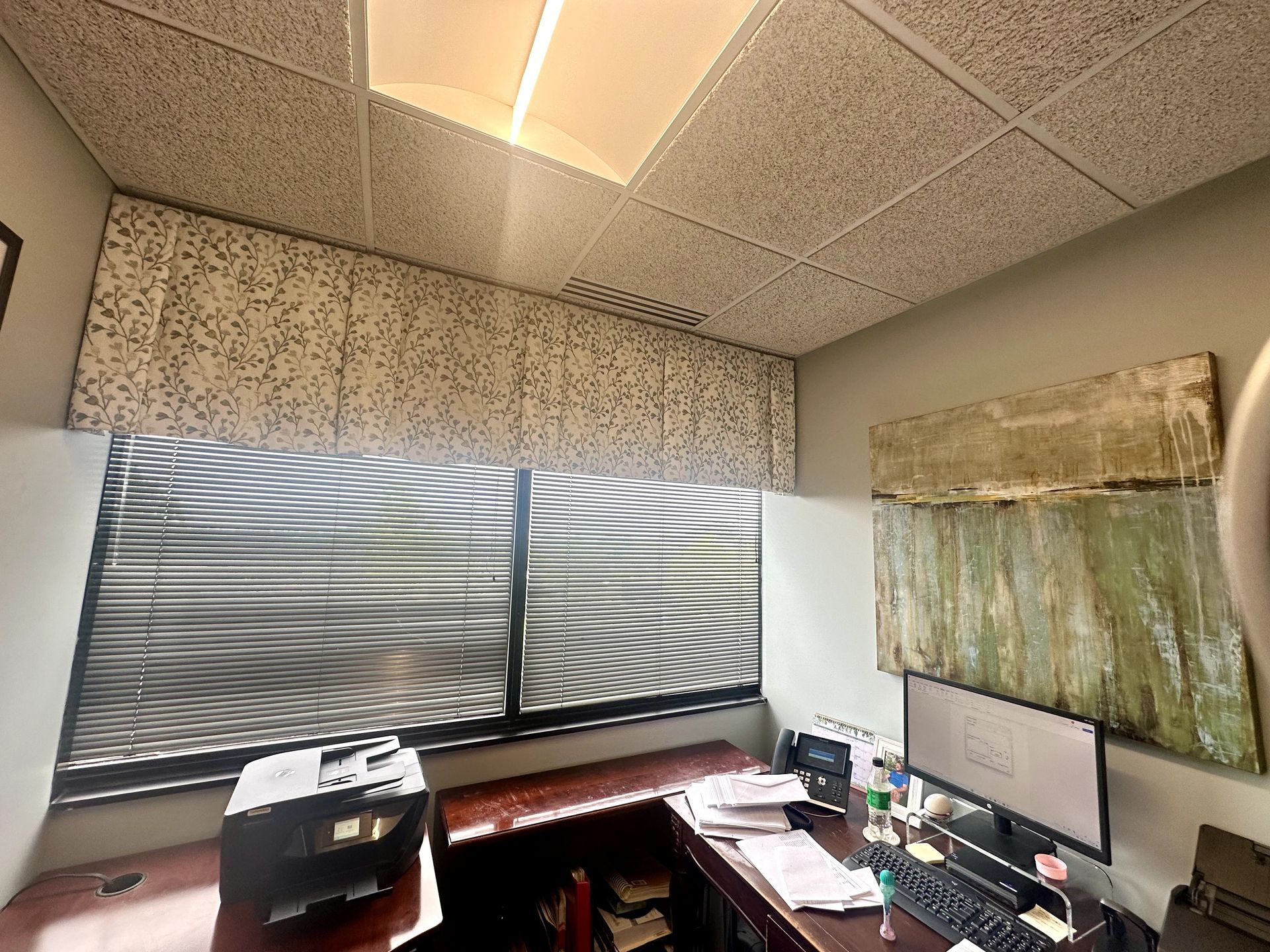 Office interior: desk with computer, printer, window with blinds, art on wall, drop ceiling, and acoustic panels.