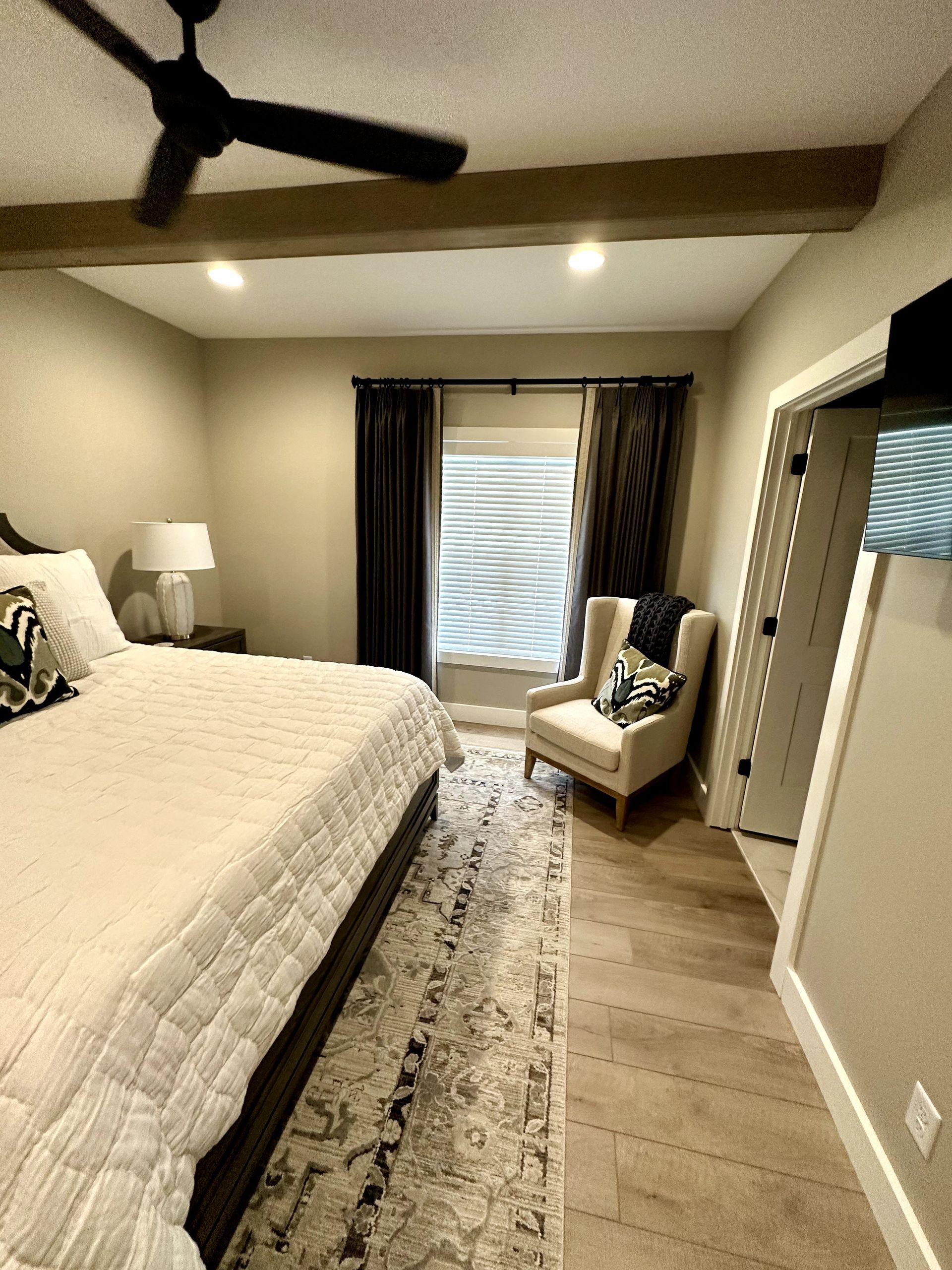 Bedroom with bed, chair, window with curtains, and a door. Beige walls and wood beams.