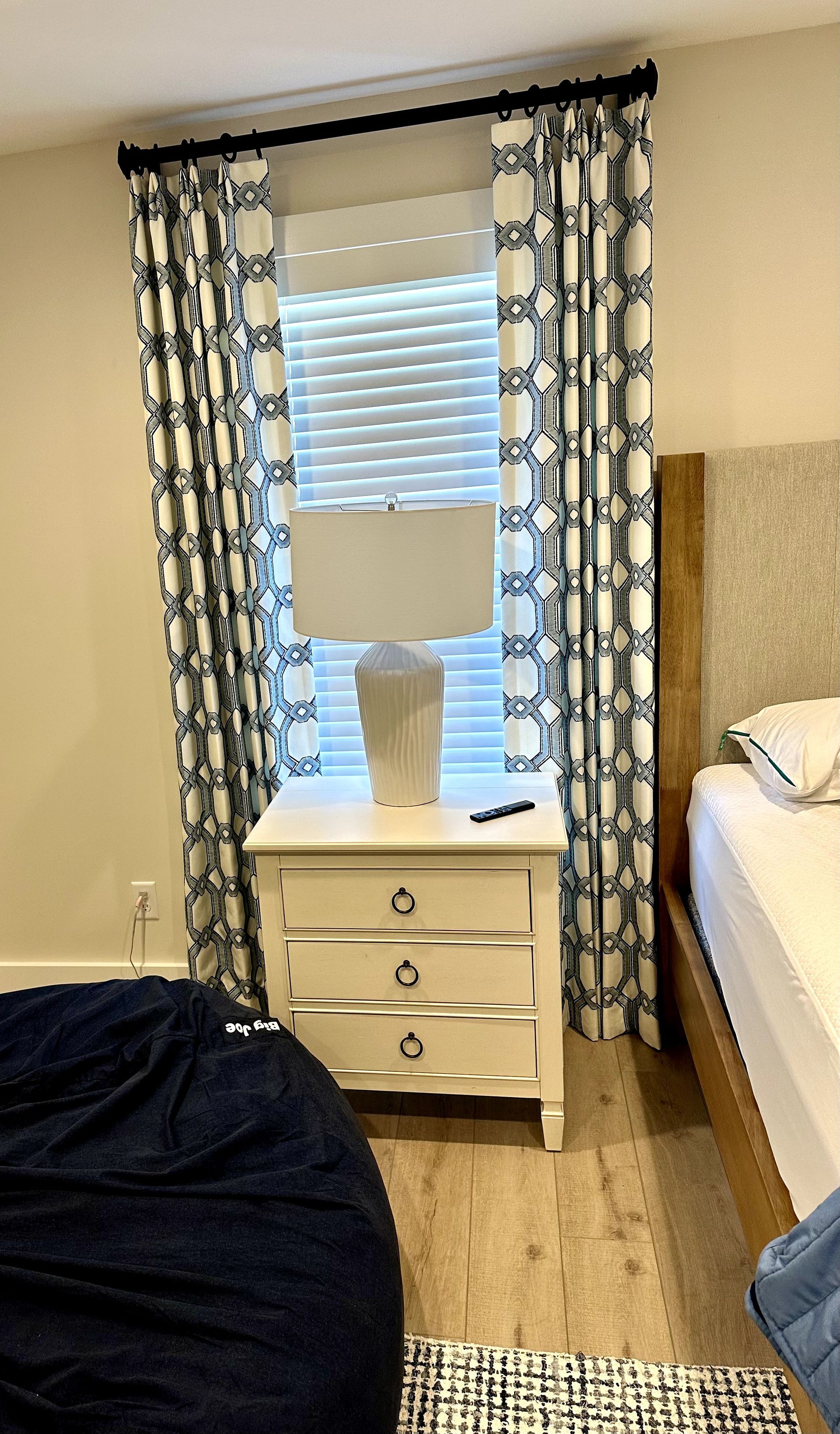 Bedroom with a white nightstand, lamp, patterned curtains, and a bed.