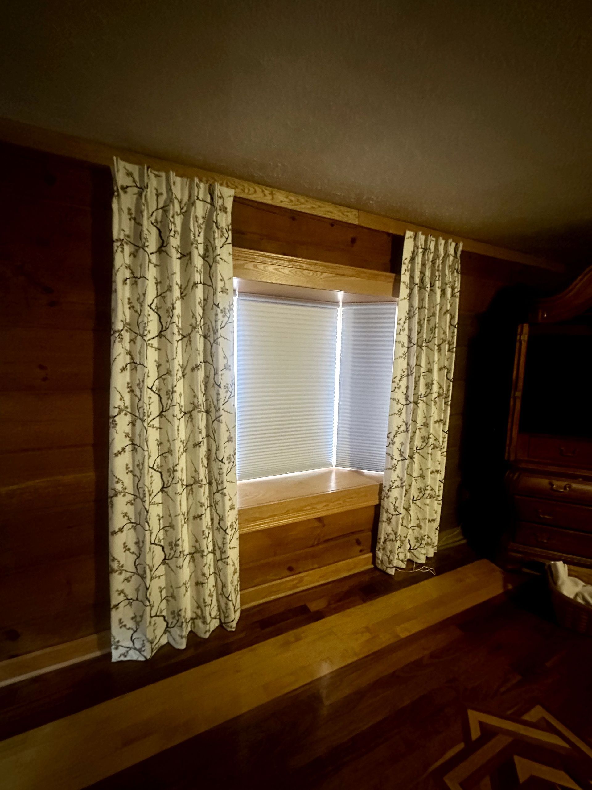 Window with floral curtains in a wood-paneled room; blinds are partially visible.