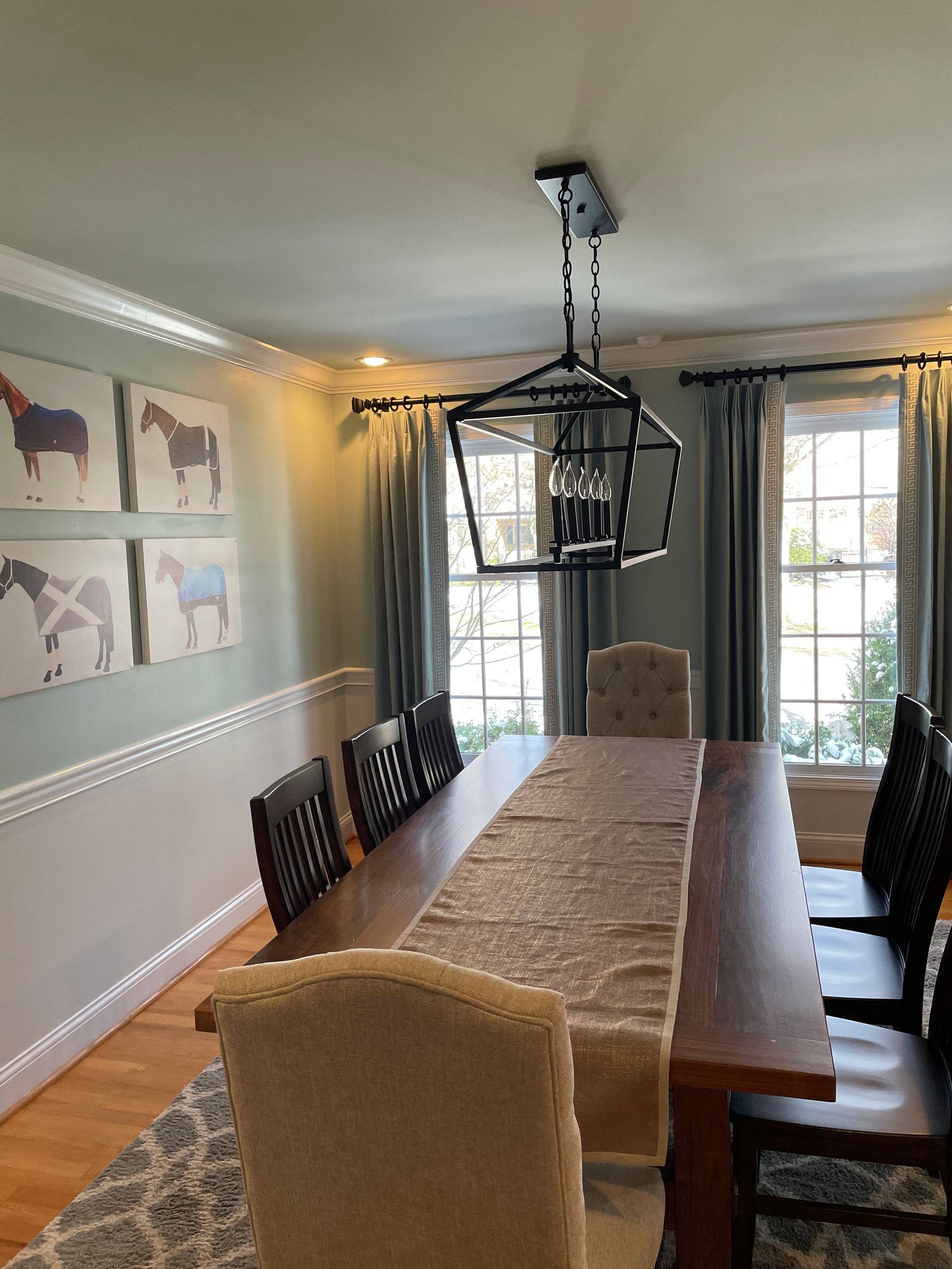 Dining room with a long wooden table, chairs, and horse paintings on the wall; light blue curtains.