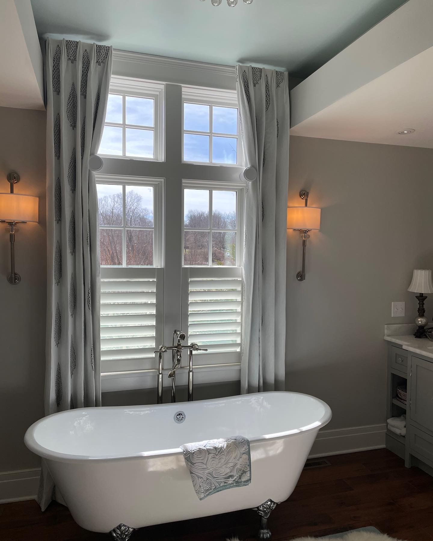Claw-foot bathtub in a gray-walled bathroom with large window, shutters, and patterned curtains.