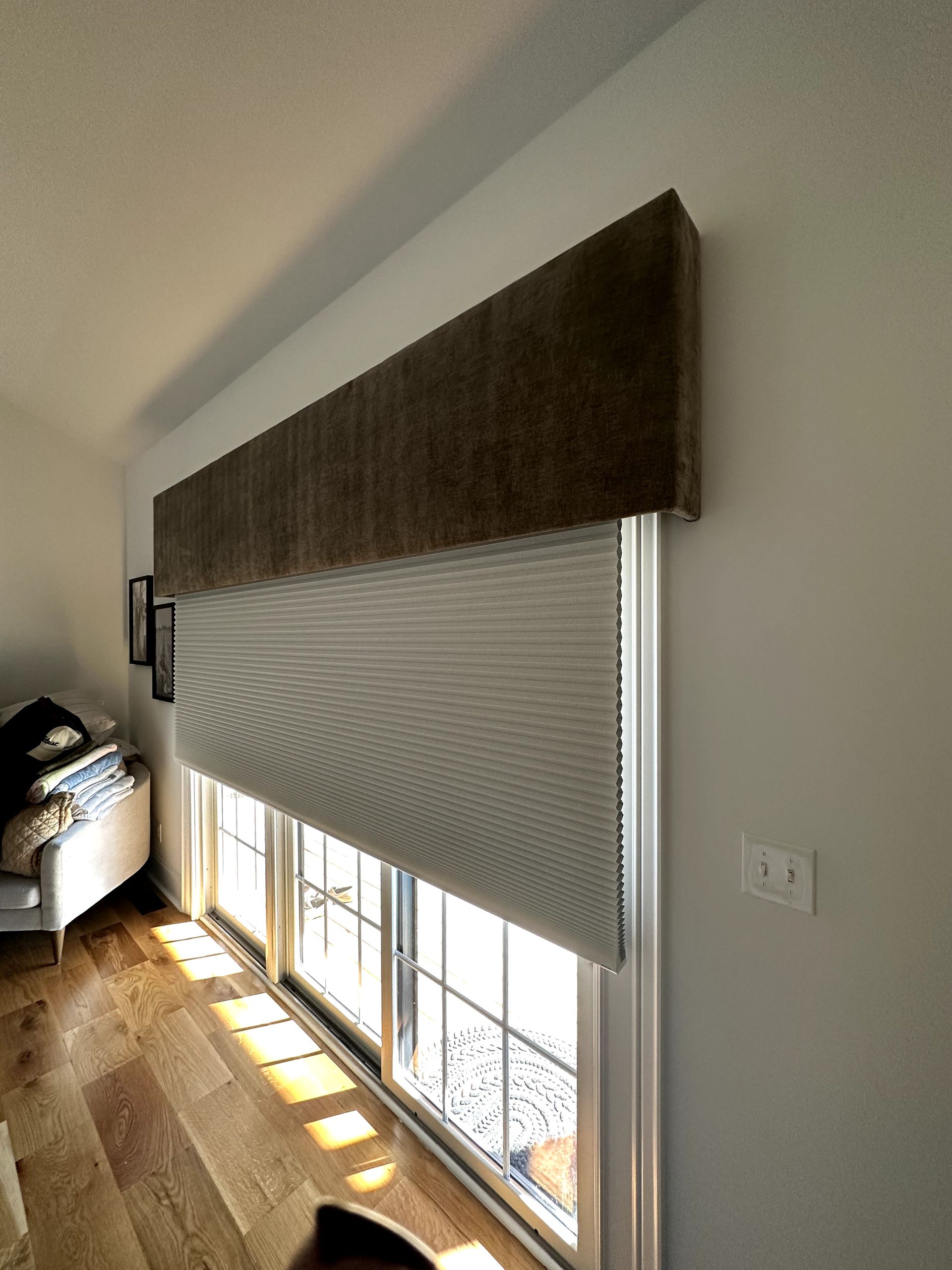 Brown upholstered valance over white cellular shades in front of a window. Sunlight streams in over hardwood floors.