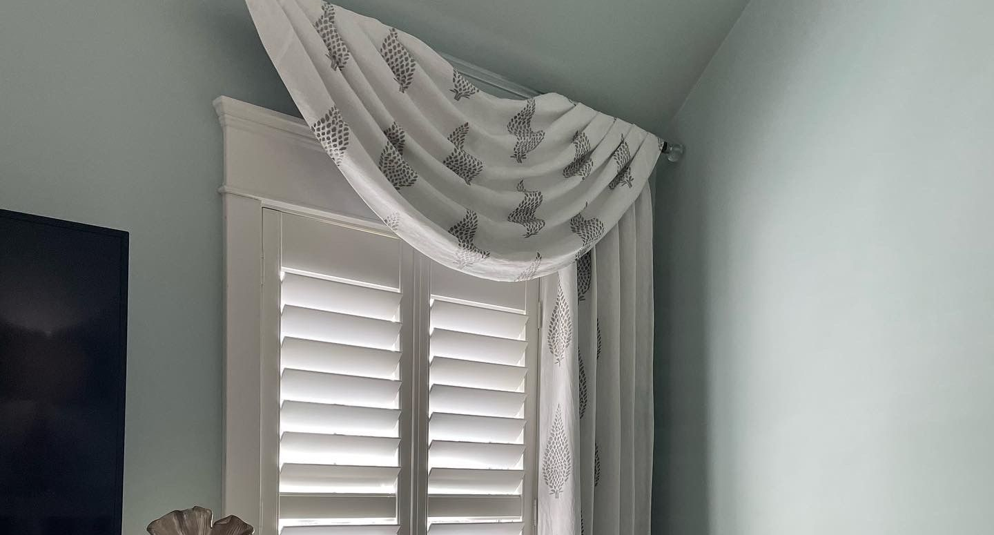 White shutters and a draped valance over a window with light blue walls.