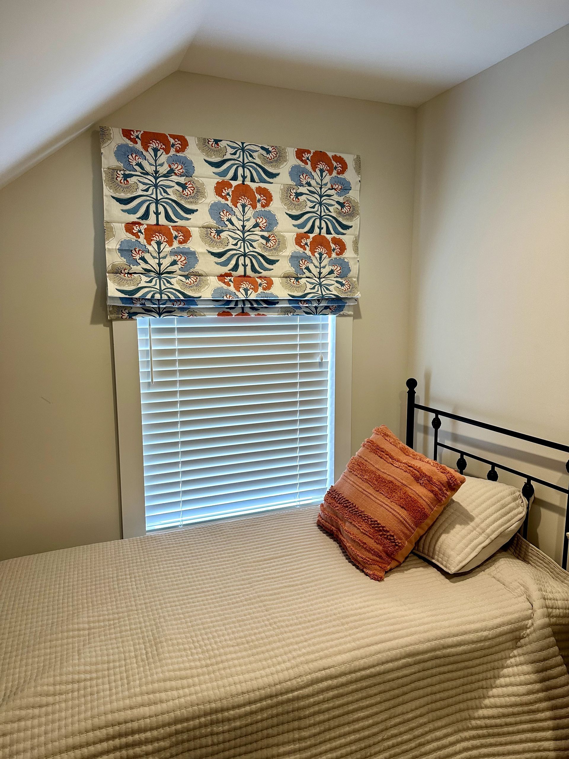 Bedroom with a twin bed, patterned Roman shade, and white blinds.  Two pillows and a white coverlet on the bed.