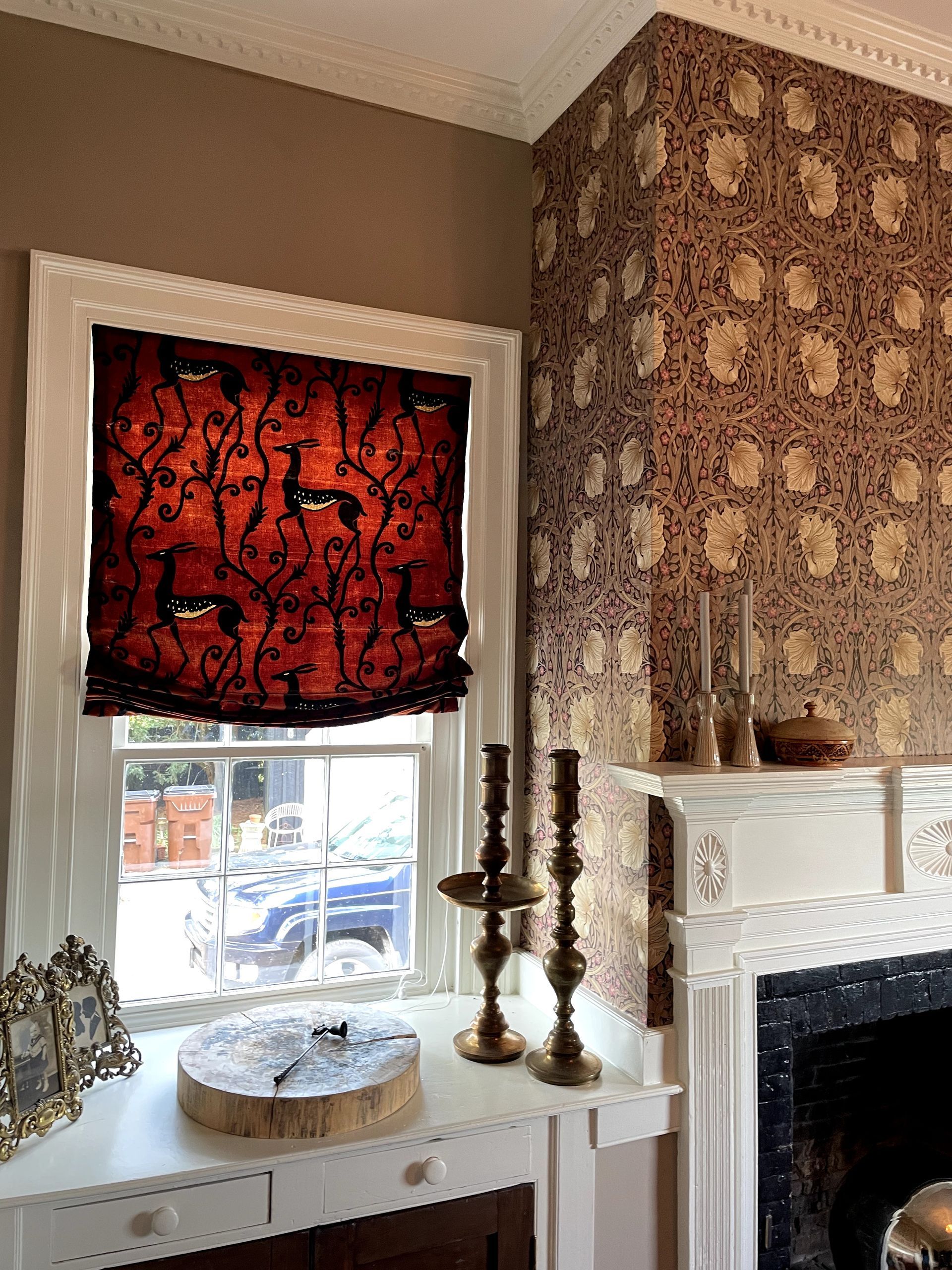 Window with patterned red shade and ornate wallpaper in a room with a white mantelpiece and candles.