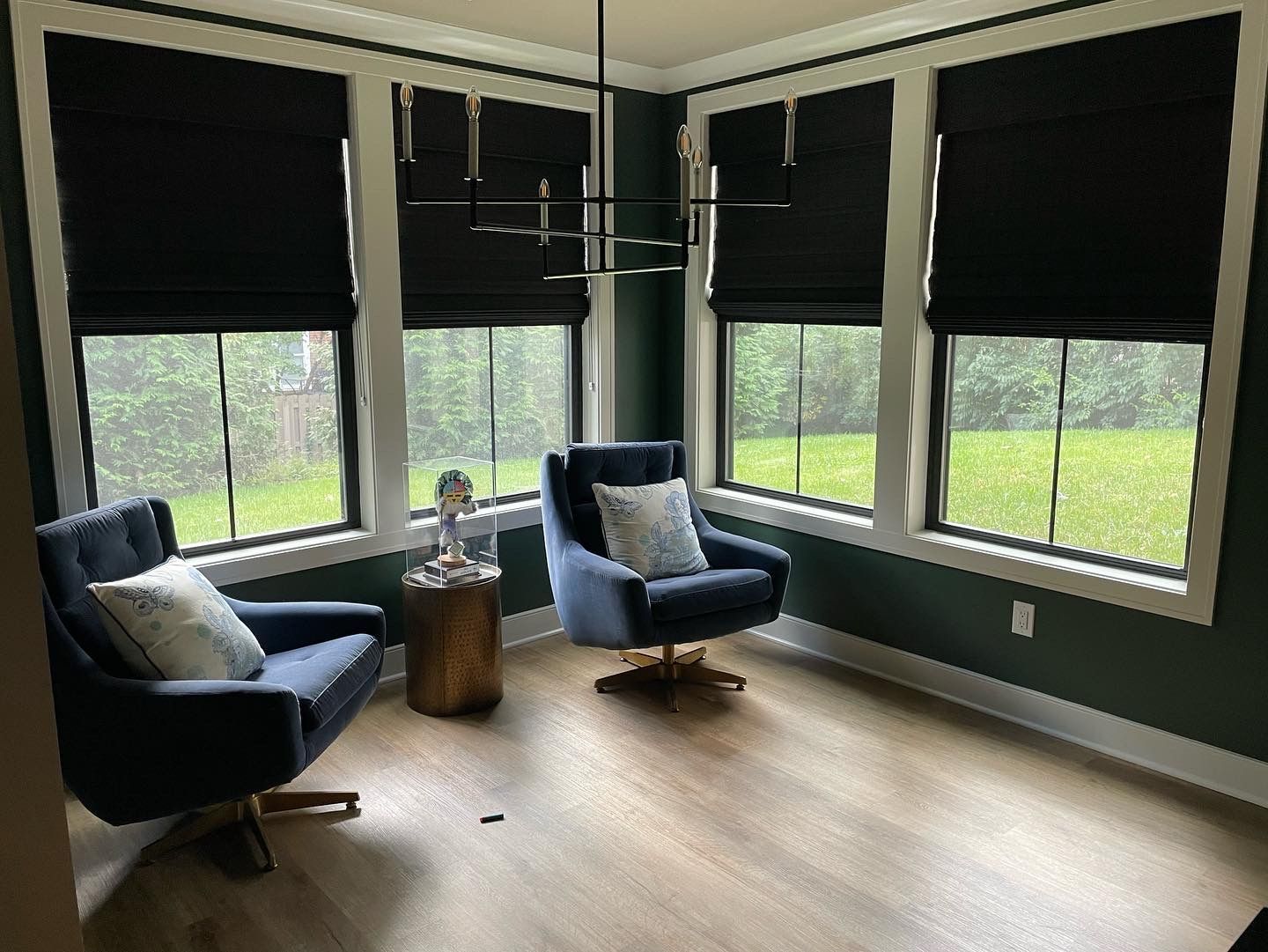 Cozy room with blue swivel chairs, black window shades, and a modern chandelier. Windows offer a view of green grass.