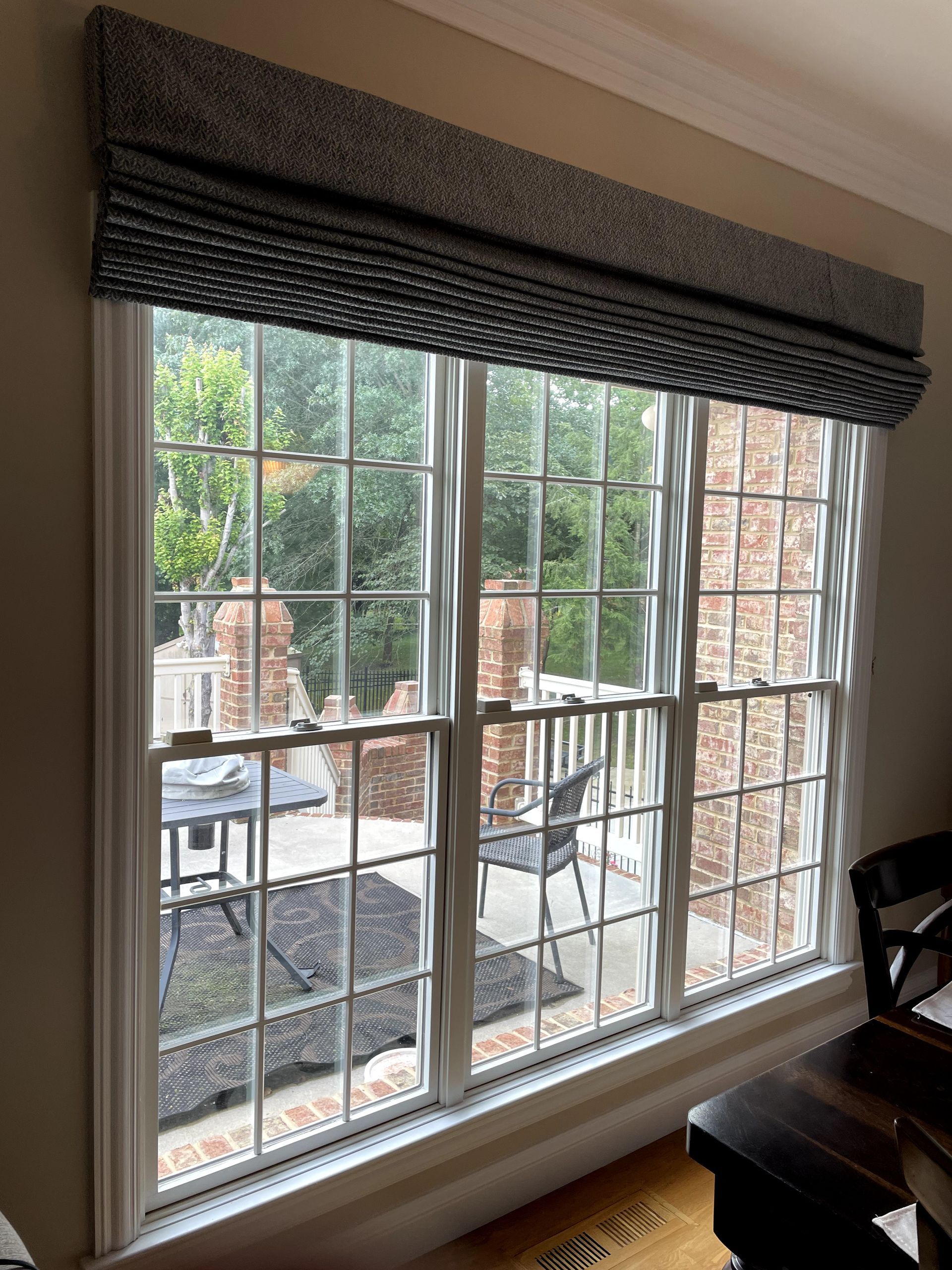 Three-paneled window with a patterned Roman shade above, overlooking a patio with seating and greenery.
