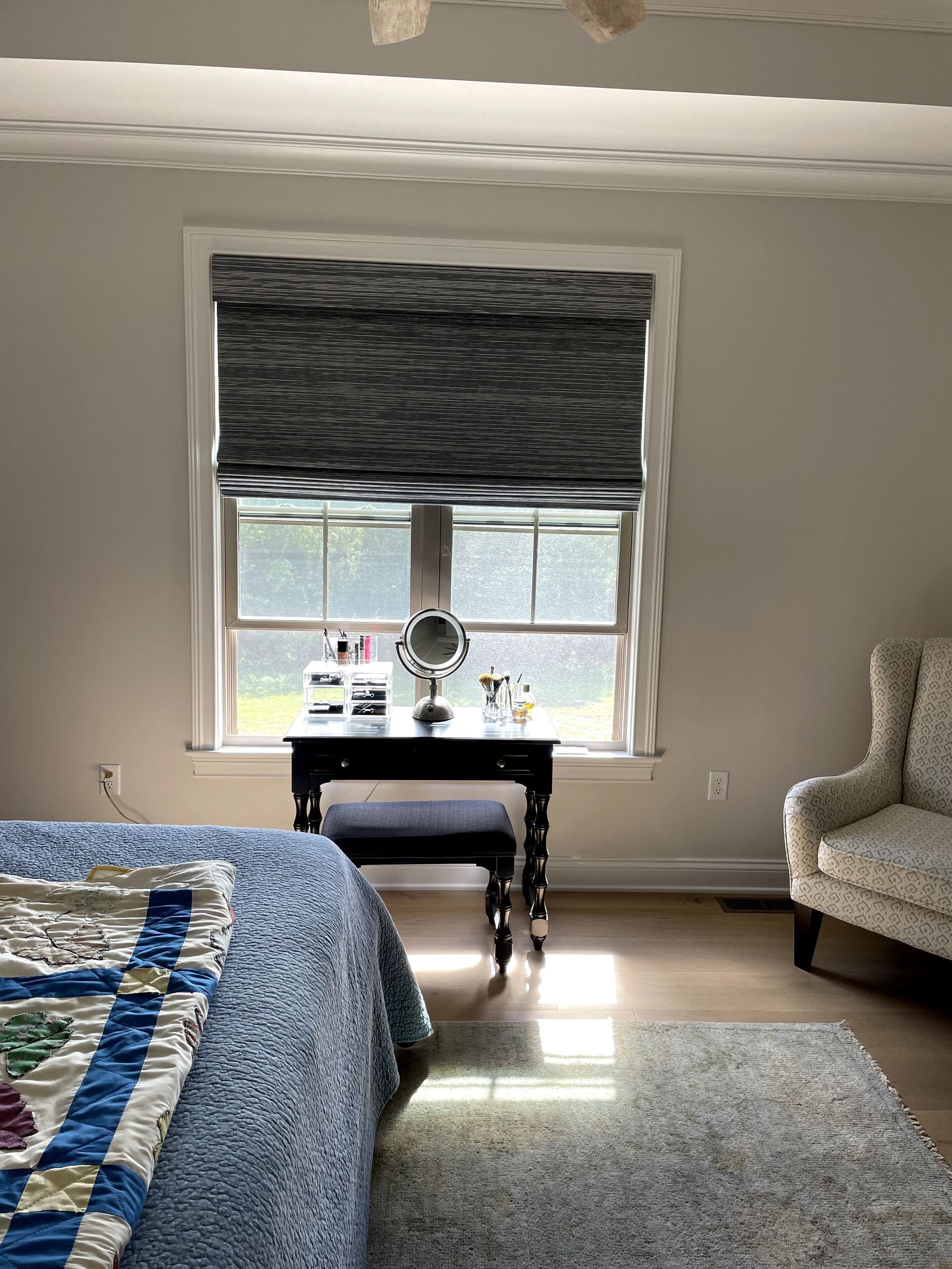 Bedroom with window, vanity, and bed. Dark patterned roman shade, light walls, and a blue quilt.