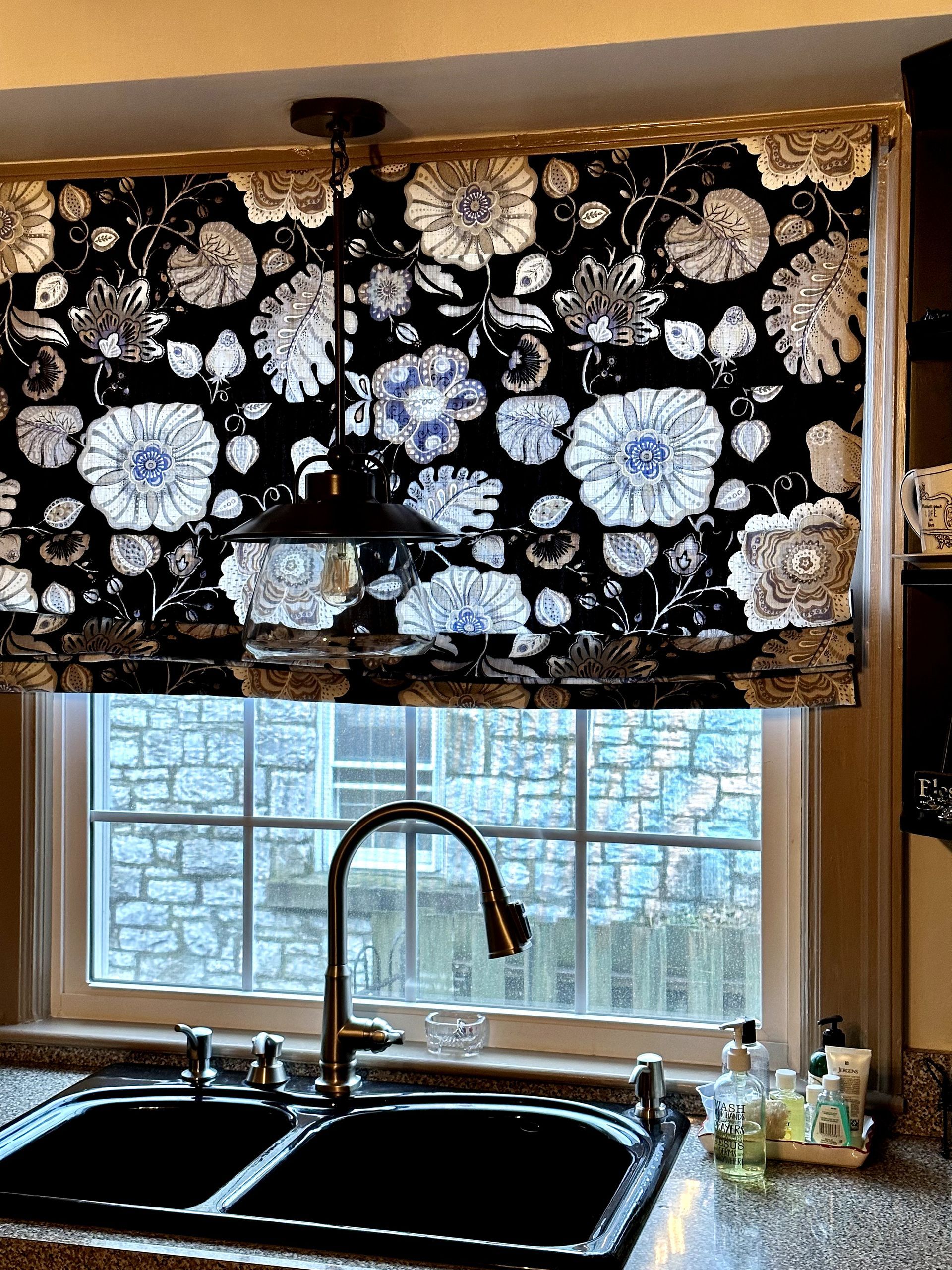 black roman shade with blue and grey flowers on the shade over a window at a sink silver faucet black painted cabinets