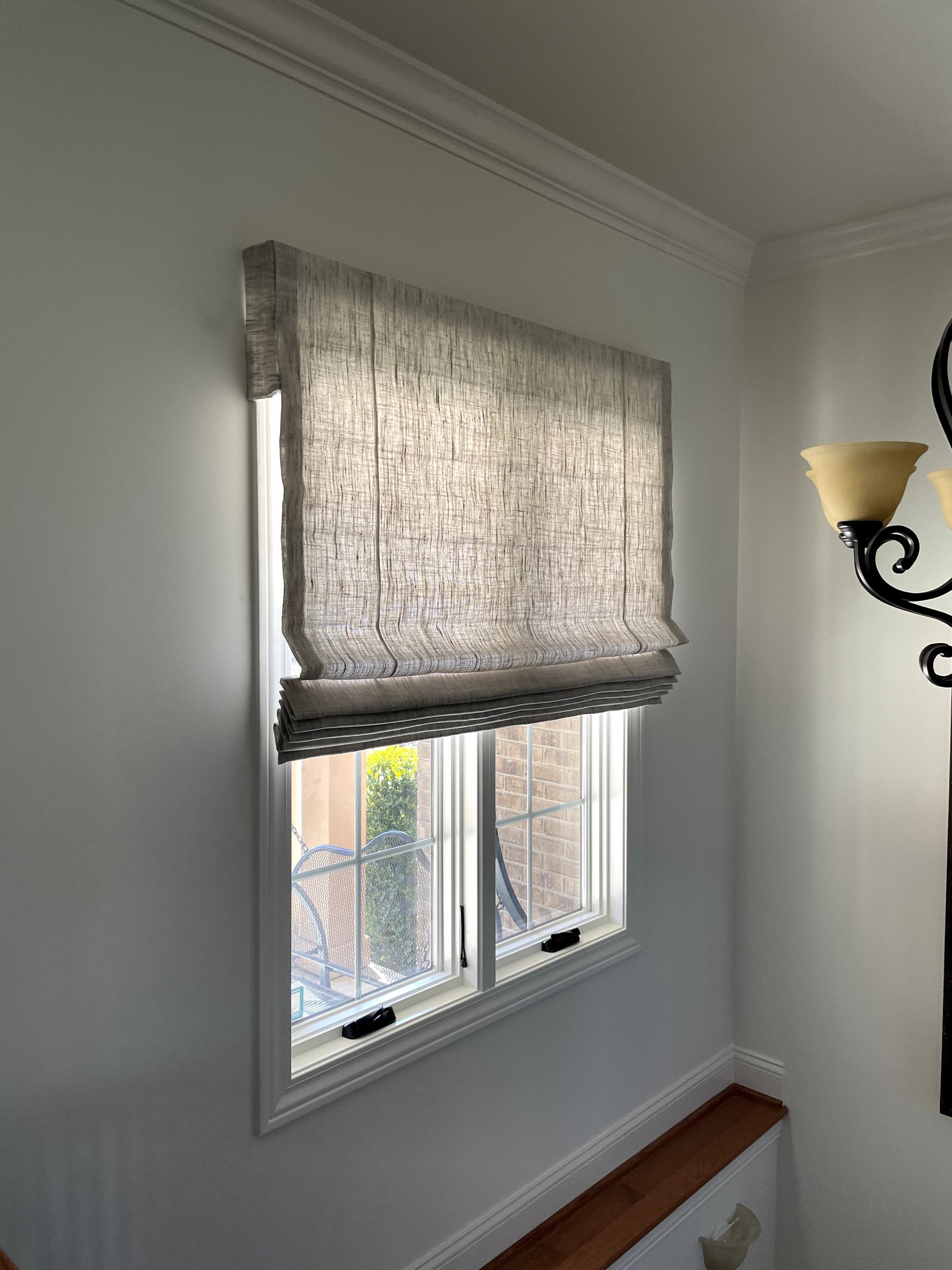 Roman shade, partially lowered, covering a window on a white wall. Brown trim, light filtering fabric.
