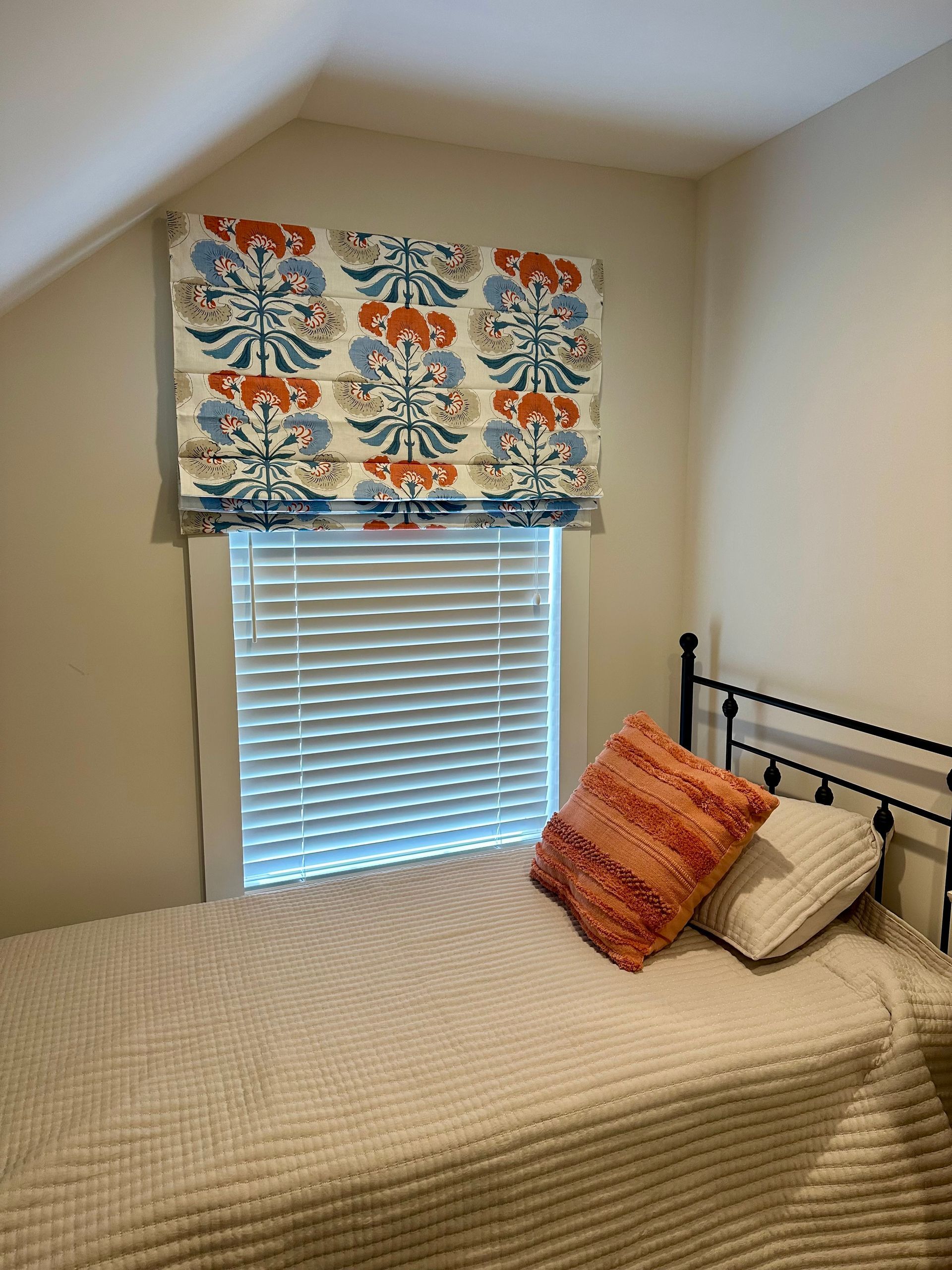 Bedroom with bed and window. Decorative floral roman shade above window with white blinds. Two pillows on the bed.