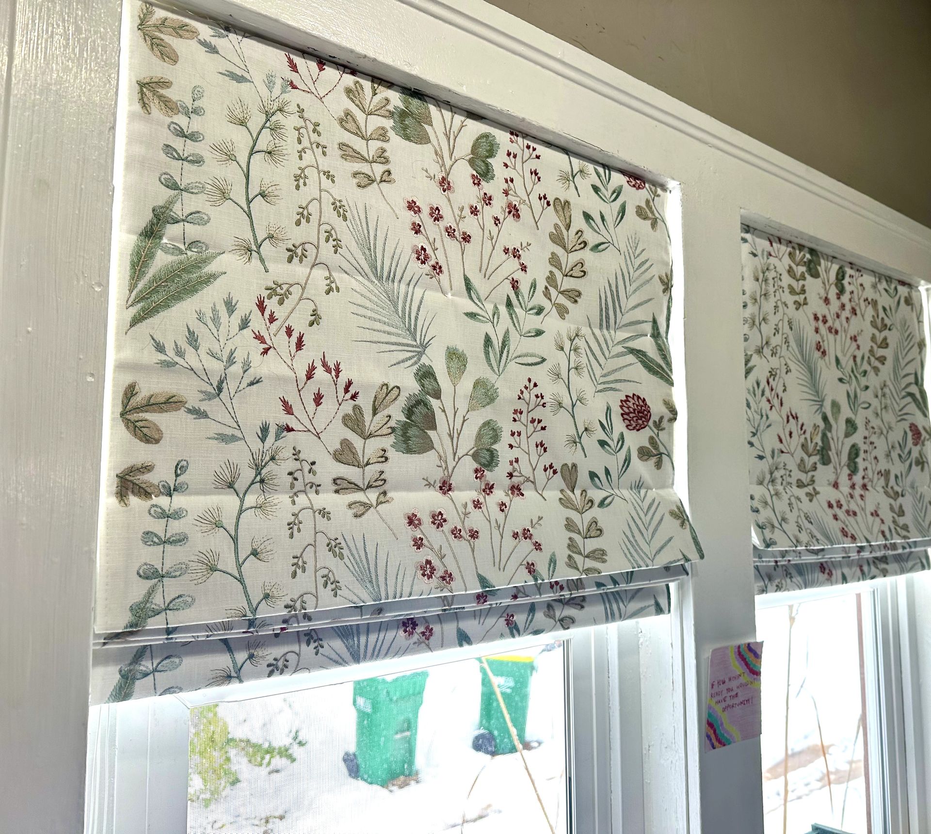Two white-framed windows with floral print Roman shades, partially raised, overlooking a scene with drawings.