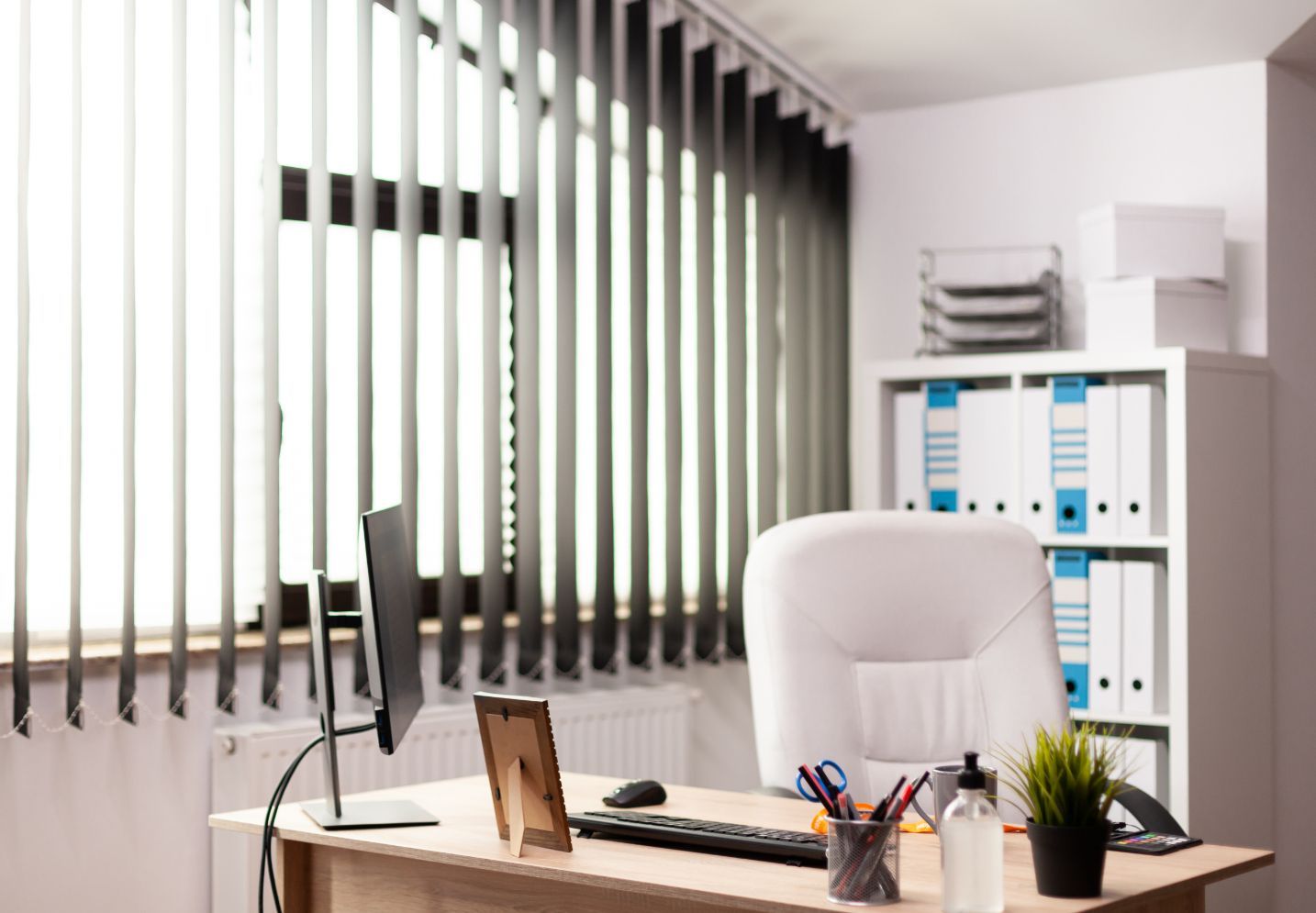 Office desk with computer, chair, and organized shelving near a window with vertical blinds.