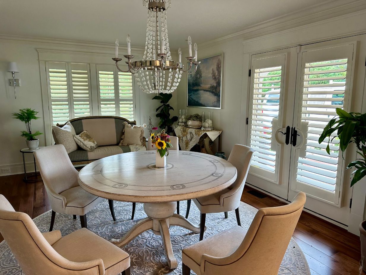 Bright dining room with round white table, beige chairs, chandelier, and large shuttered windows.
