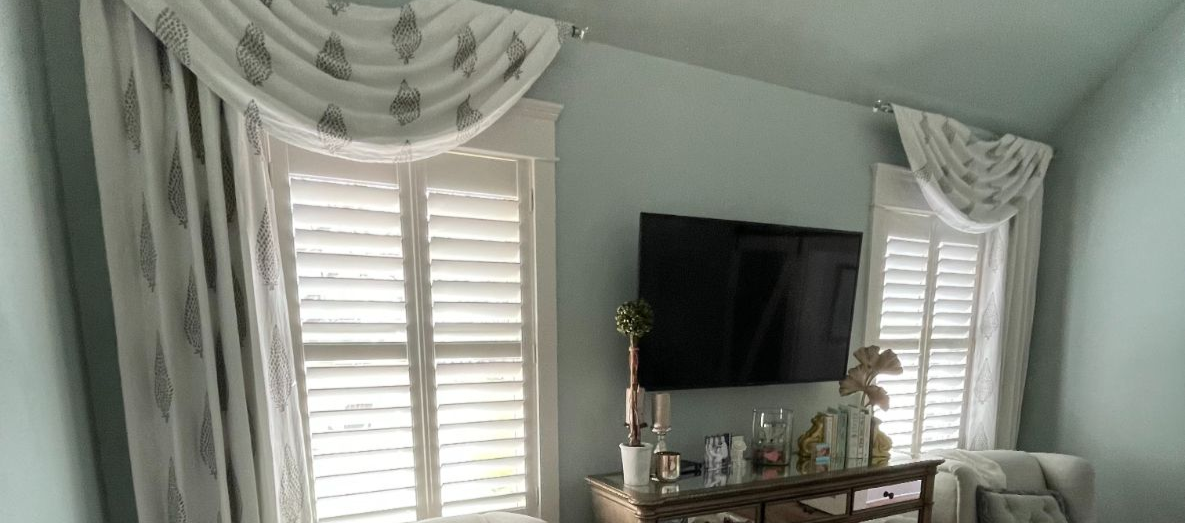 Bright living room with white shutters, patterned drapes, wall-mounted TV, and small table with decor