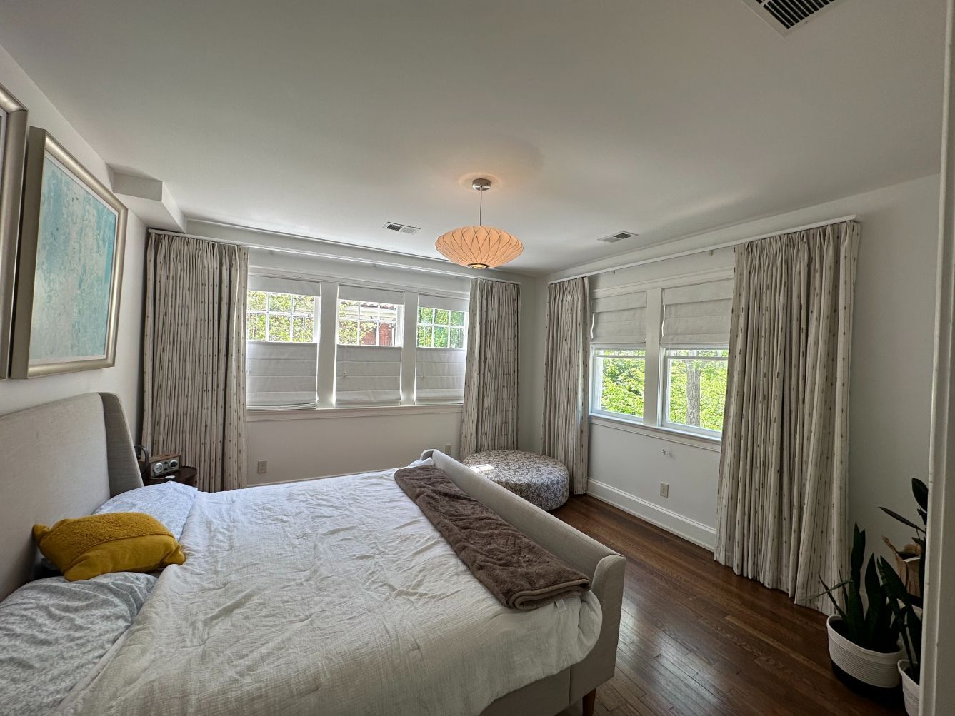 A bright bedroom with a bed, wood floors, and two large windows draped with patterned floor-length curtains.