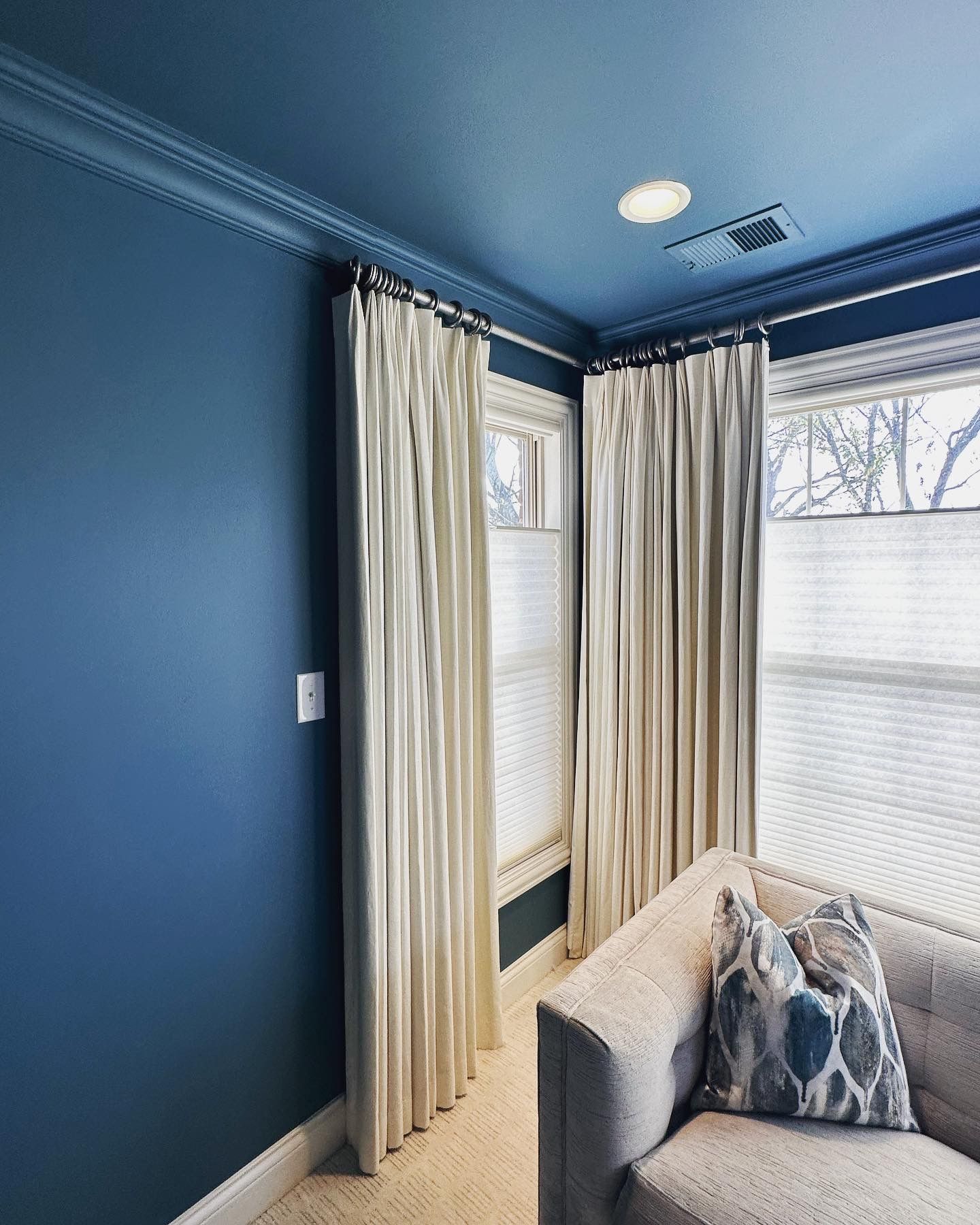 Blue-walled room with cream drapes framing windows. Beige sofa with patterned pillow.