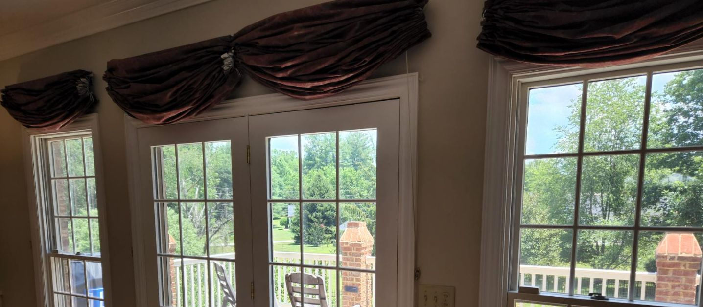 Three tall windows with dark drapes overlooking a bright garden and brick fence