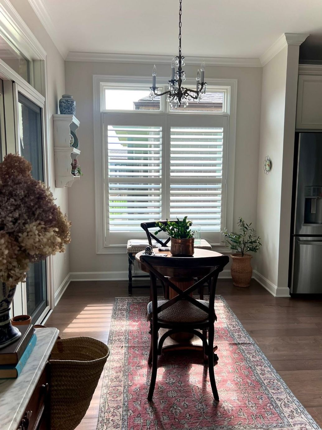 Dining area with table, chairs, rug, and window with shutters; light streams in.