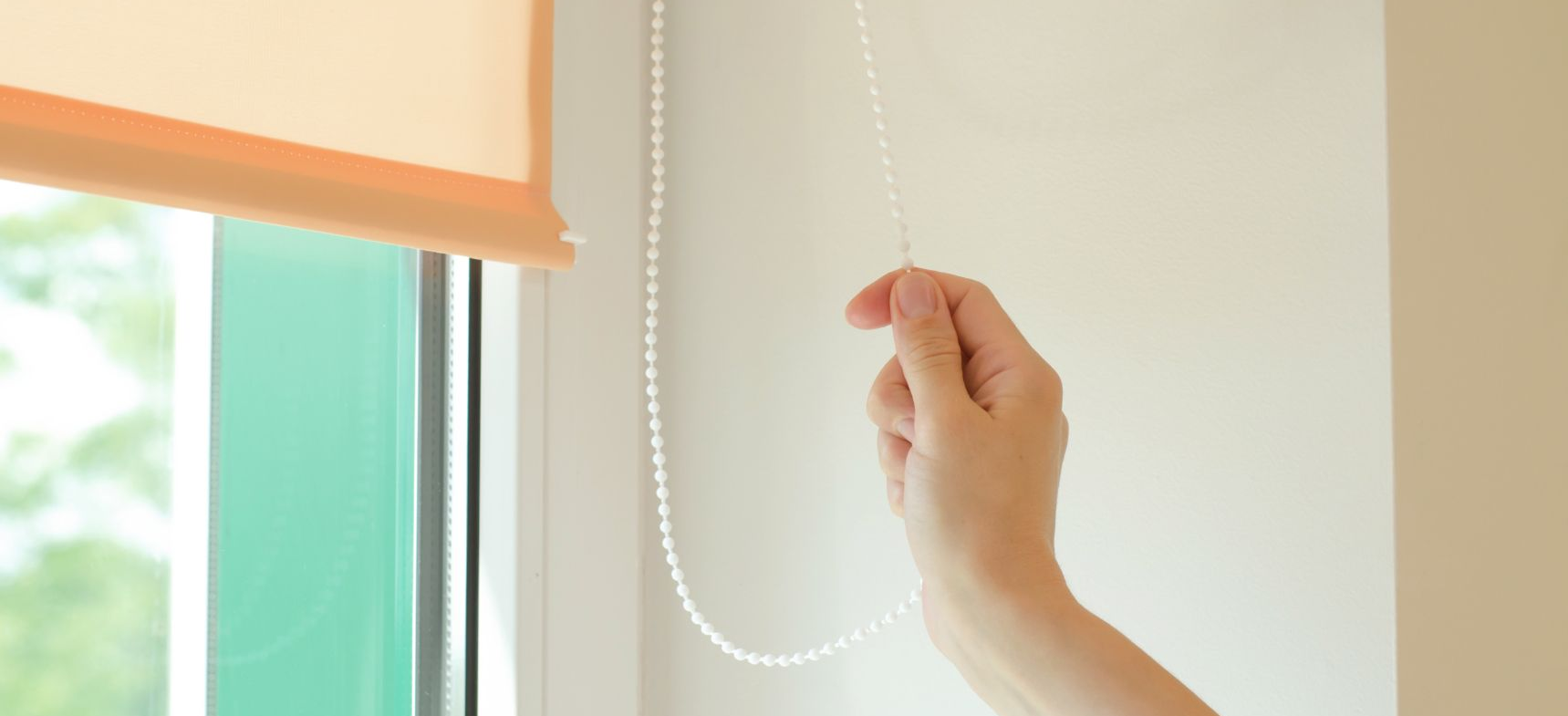 A hand pulls a beaded chain to adjust a window shade.