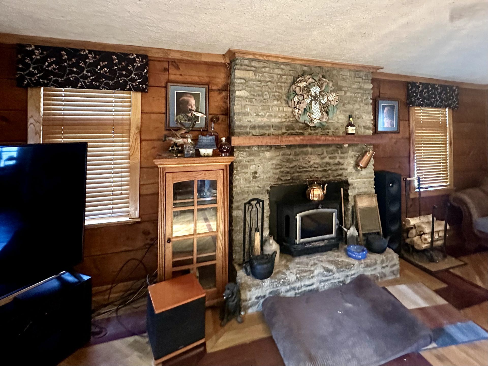 Living room with stone fireplace, wood paneling, and windows with blinds.