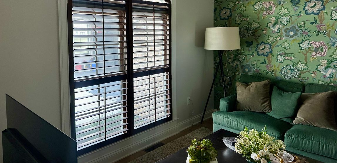 A room with a window with shutters, green couch and floral wallpaper.