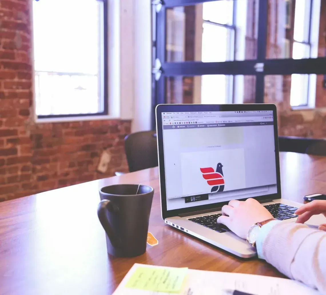 A person types on a laptop at a wooden table with a black mug, situated in an office with brick walls and large windows.