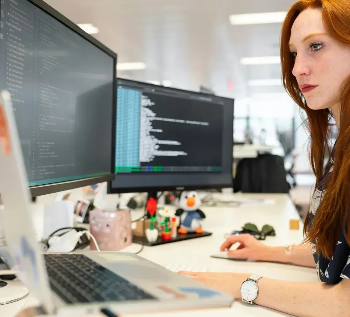 A person with long red hair focuses on code displayed across multiple monitors at a clean, professional office desk.