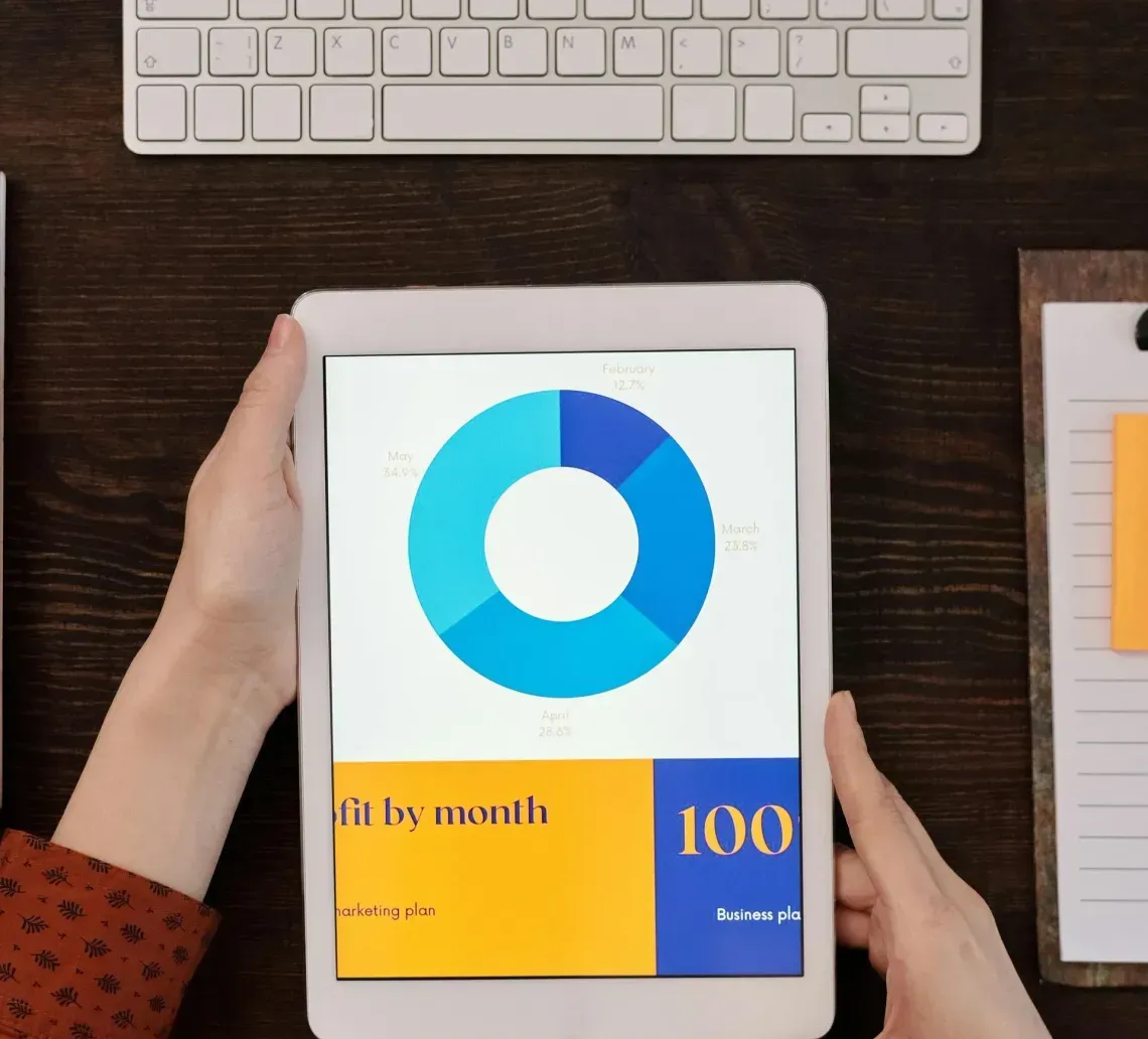 Overhead view of hands holding a tablet displaying a colorful circular chart, set on a dark desk with a keyboard.