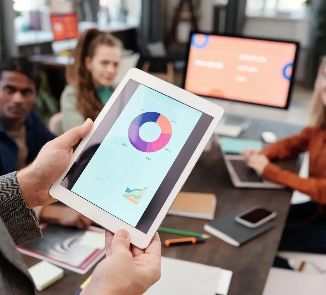 A hand holds a tablet displaying a pie chart during a team meeting in an office setting.