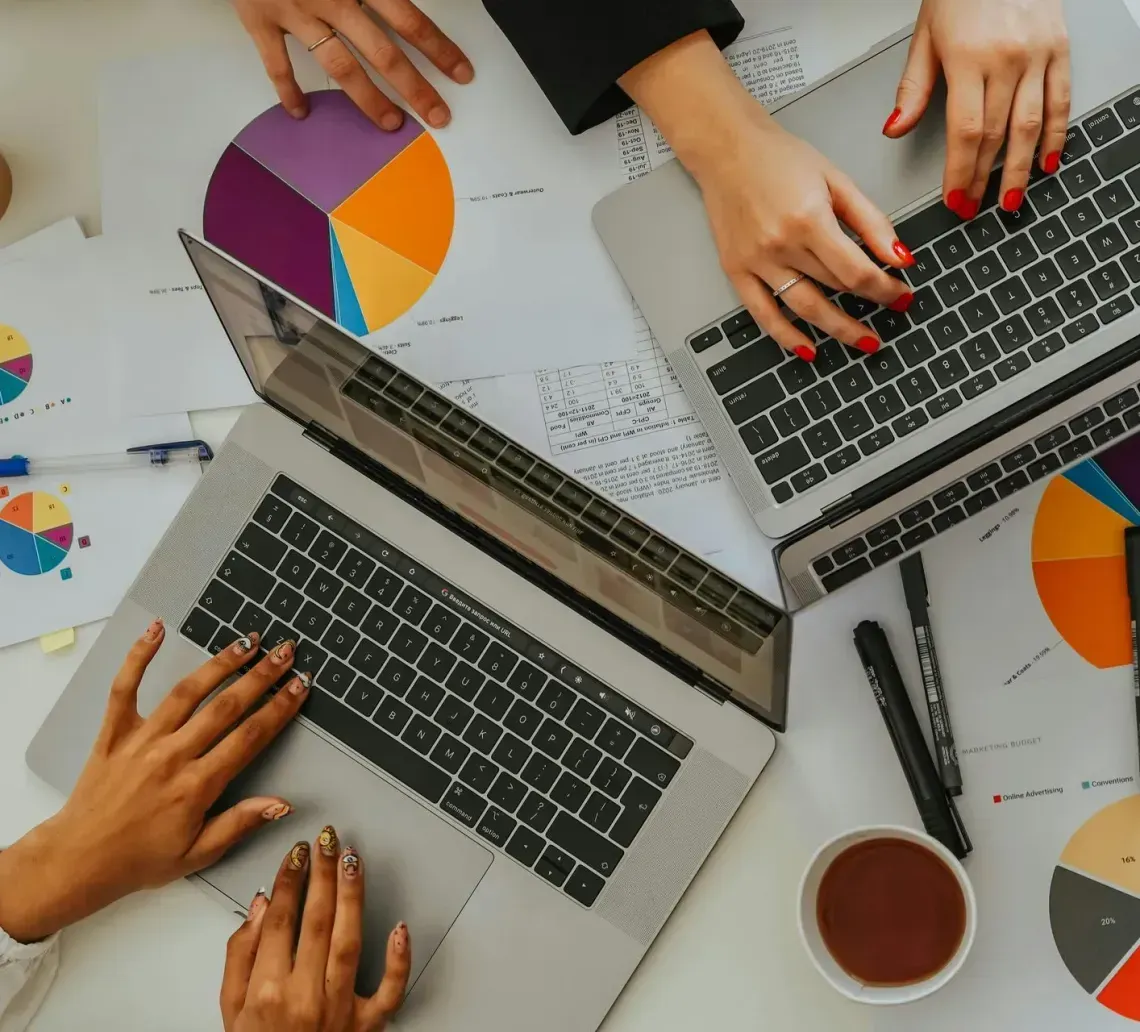 Two people use laptops on a desk covered with colorful pie charts, pens, and coffee cups.
