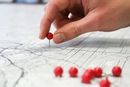 A hand places a red pin onto a map showing grid lines and contour sketches.